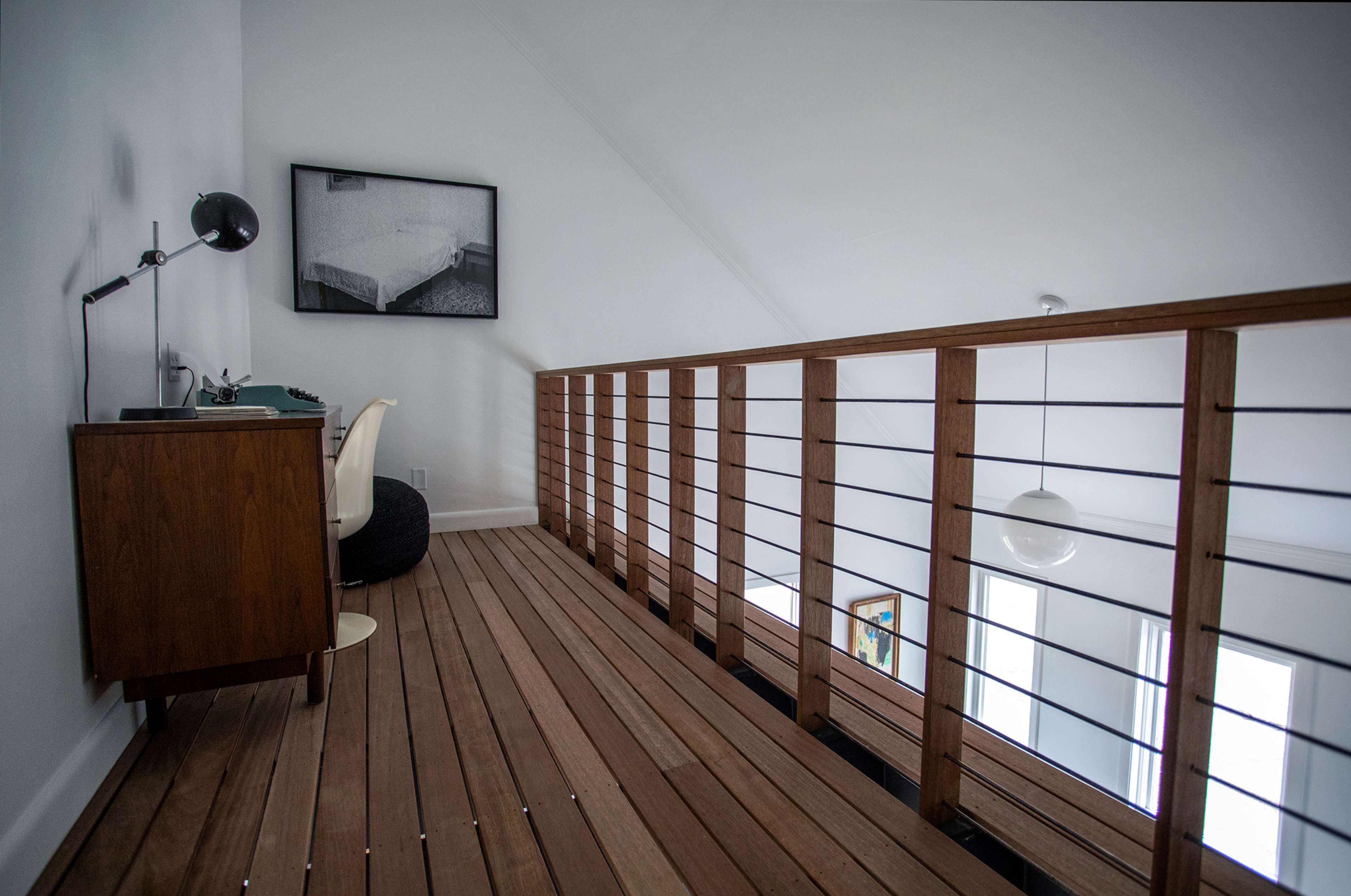 A wooden balcony features a desk lamp, a vintage typewriter, and a wooden dresser, overlooking a minimalist interior space with large windows.