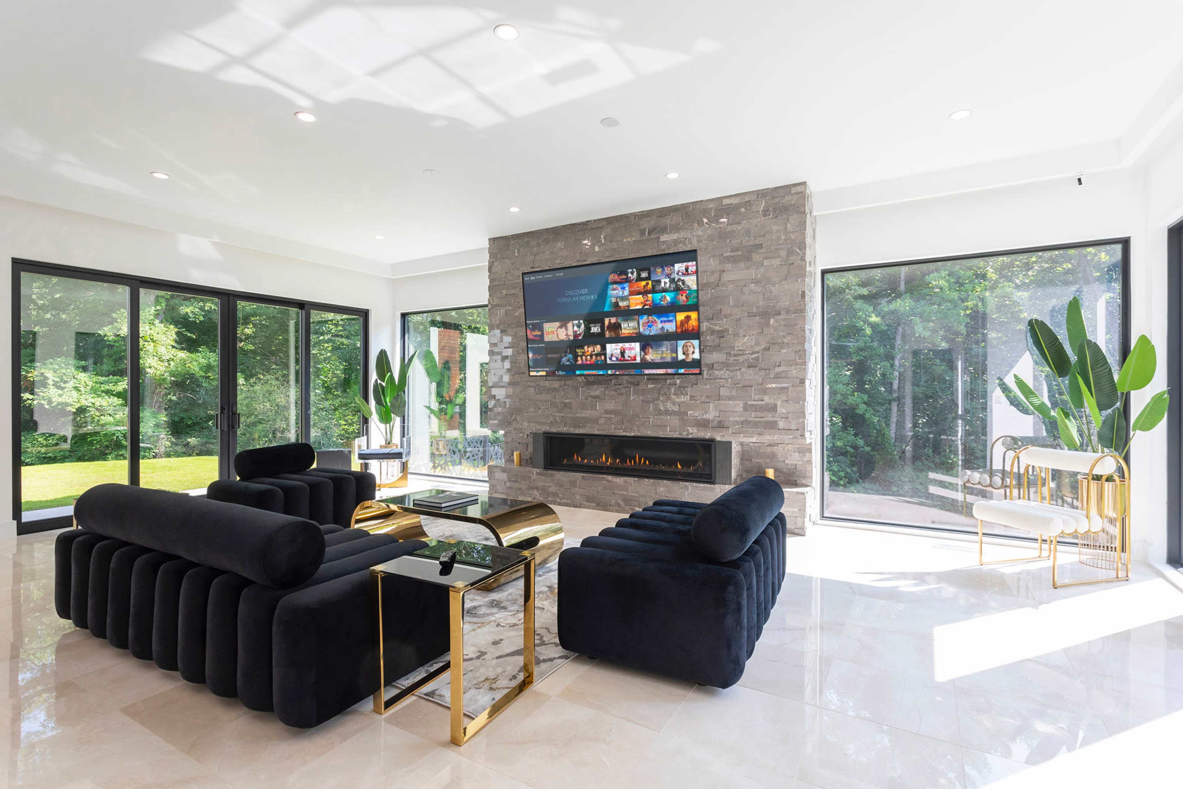 A modern living room features a stone fireplace wall with a large television above, surrounded by large windows and contemporary furniture in black and gold.