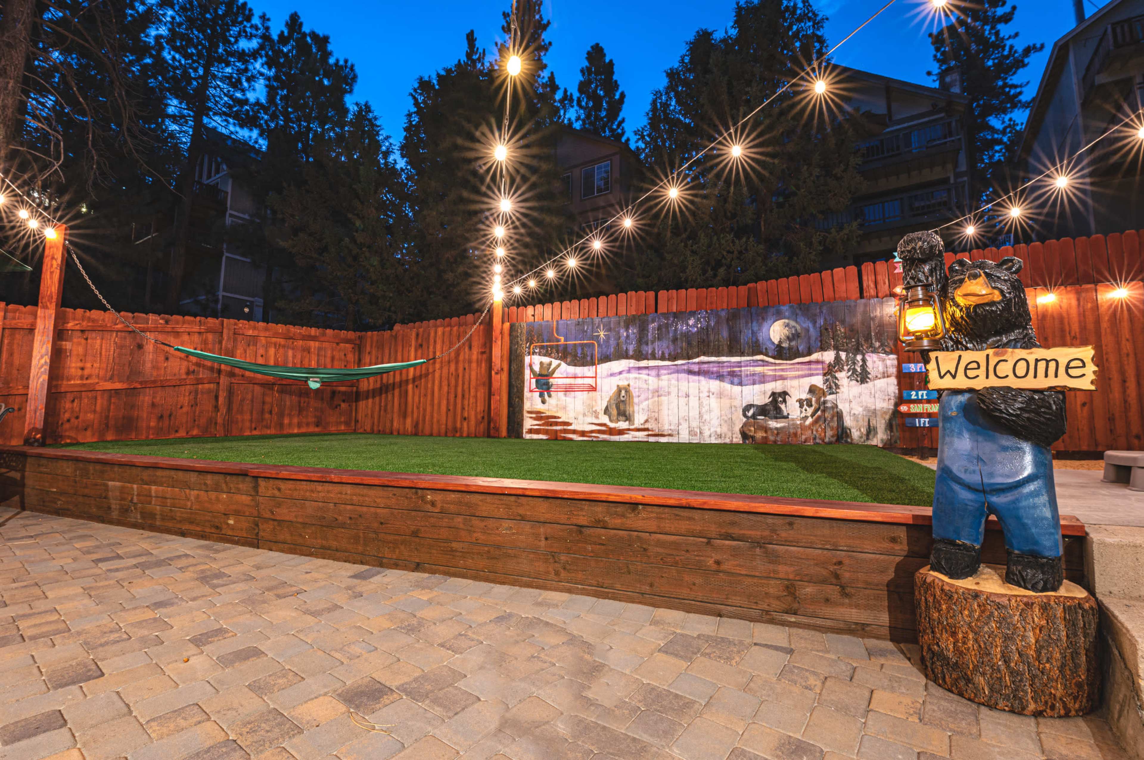 The image shows a landscaped backyard with a wooden deck, a hammock, a mural of a winter scene, string lights, and a bear statue holding a "Welcome" sign.