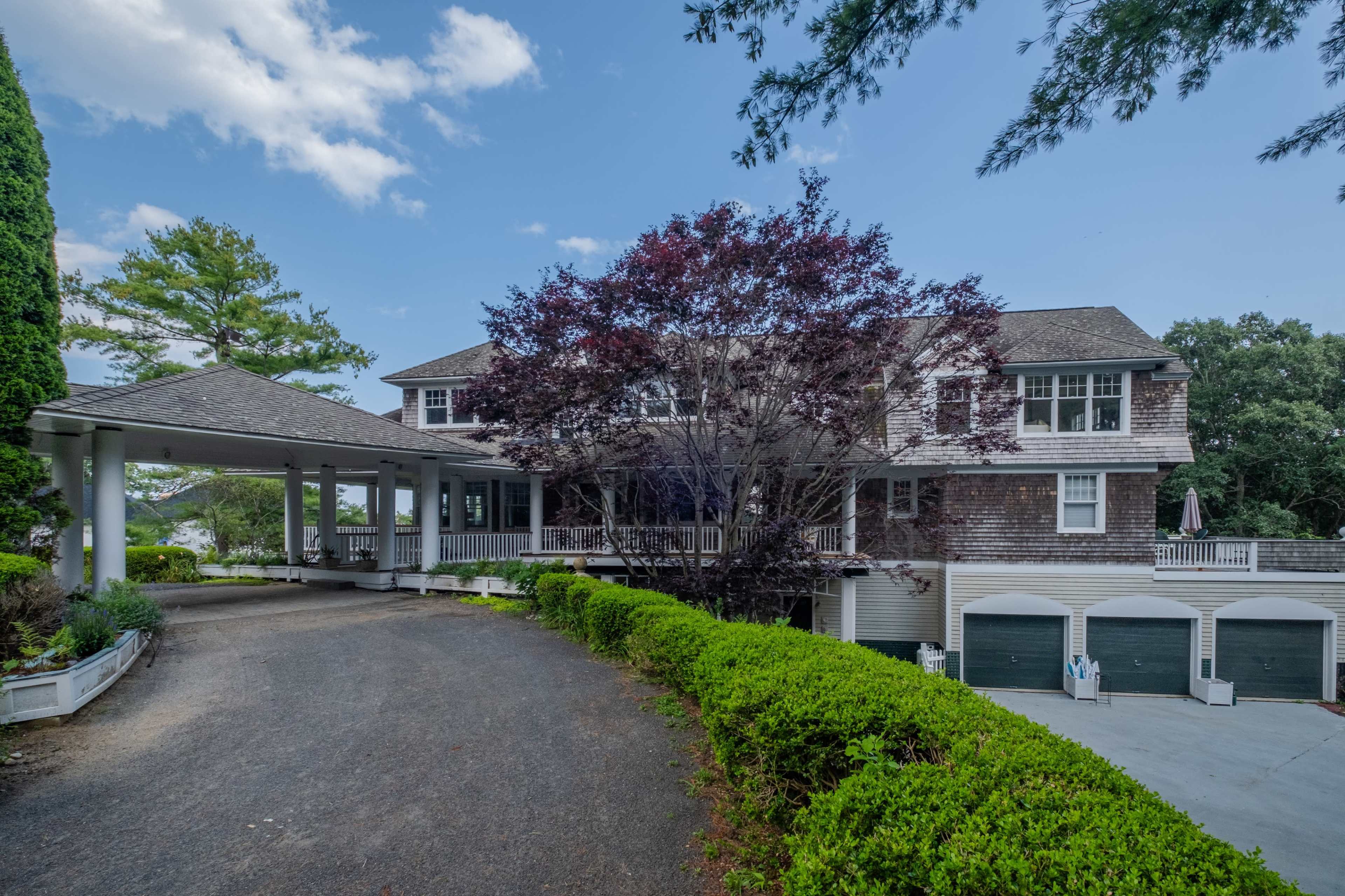 A large, multi-story house with a wraparound porch and a landscaped driveway is set against a clear blue sky.