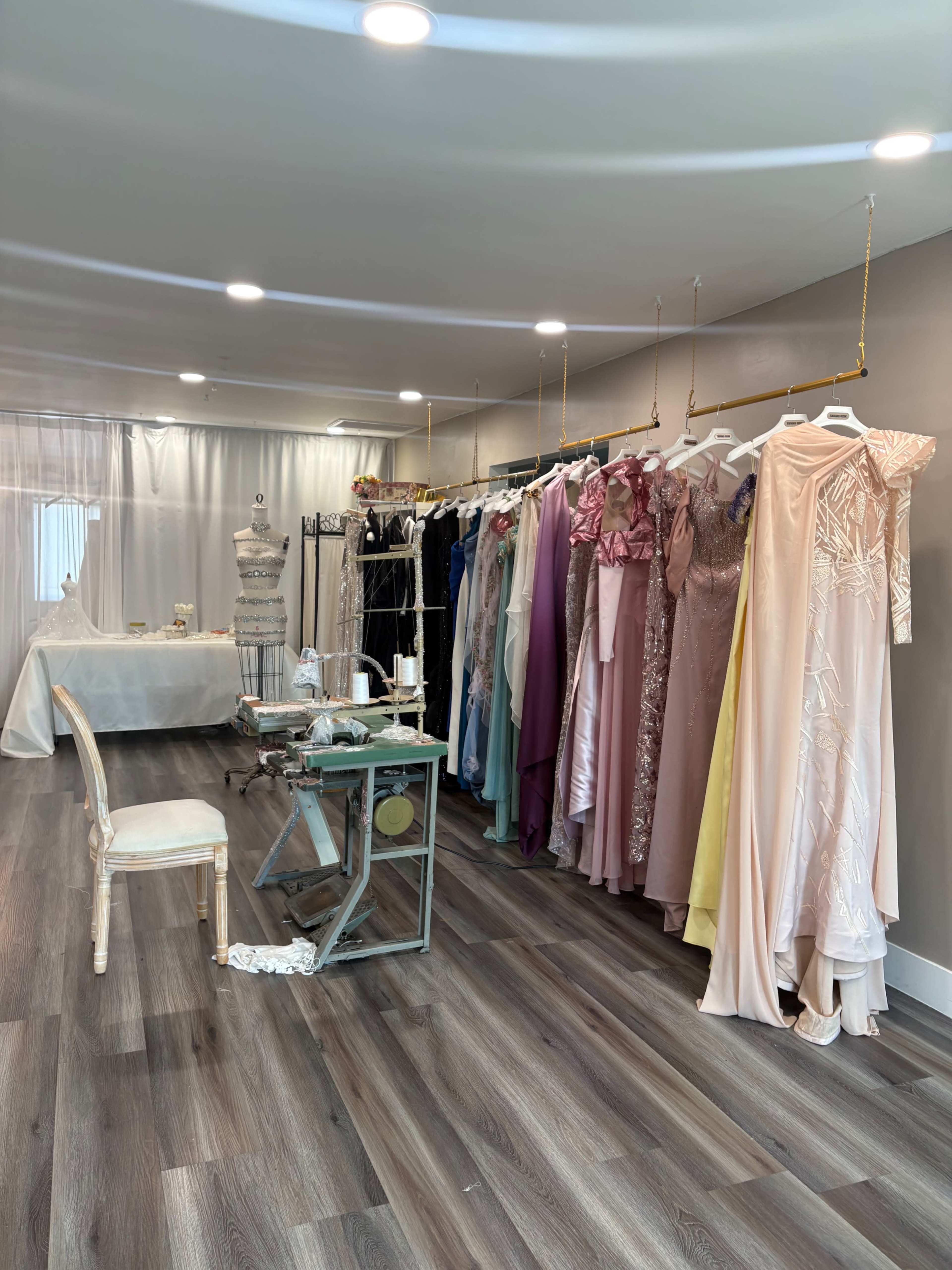 The image shows a bridal boutique interior with a variety of evening gowns displayed on hangers along one wall, a dressmaking table in the center, and a white backdrop in the background.