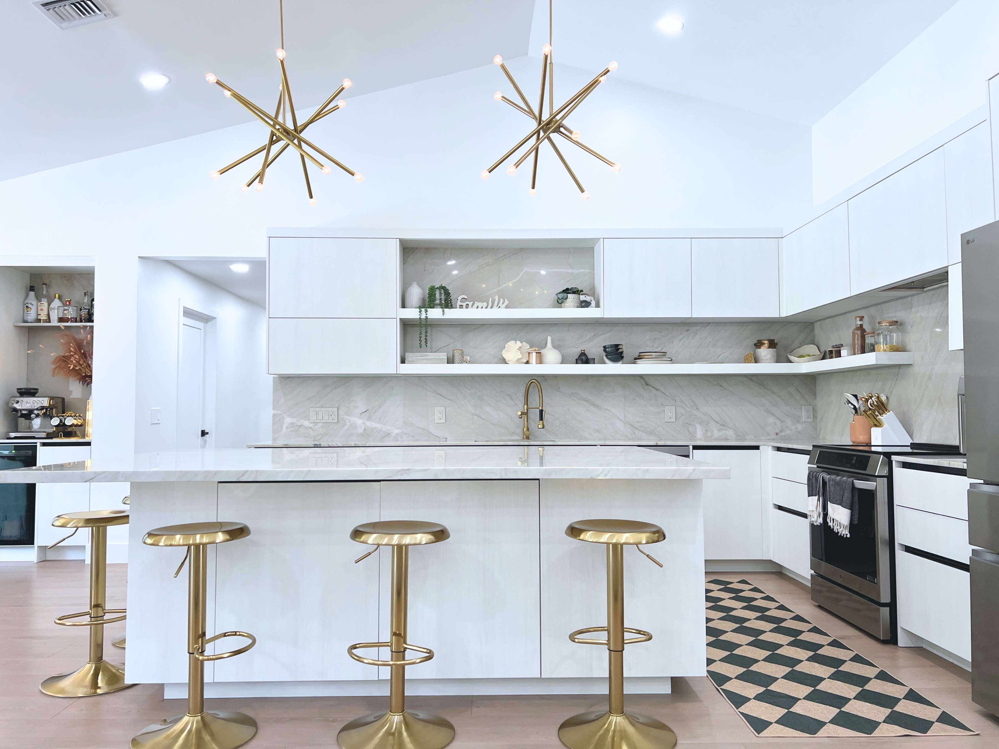 The image shows a modern kitchen featuring white cabinets, a large central island with a marble top, gold bar stools, and two striking light fixtures hanging from the ceiling.