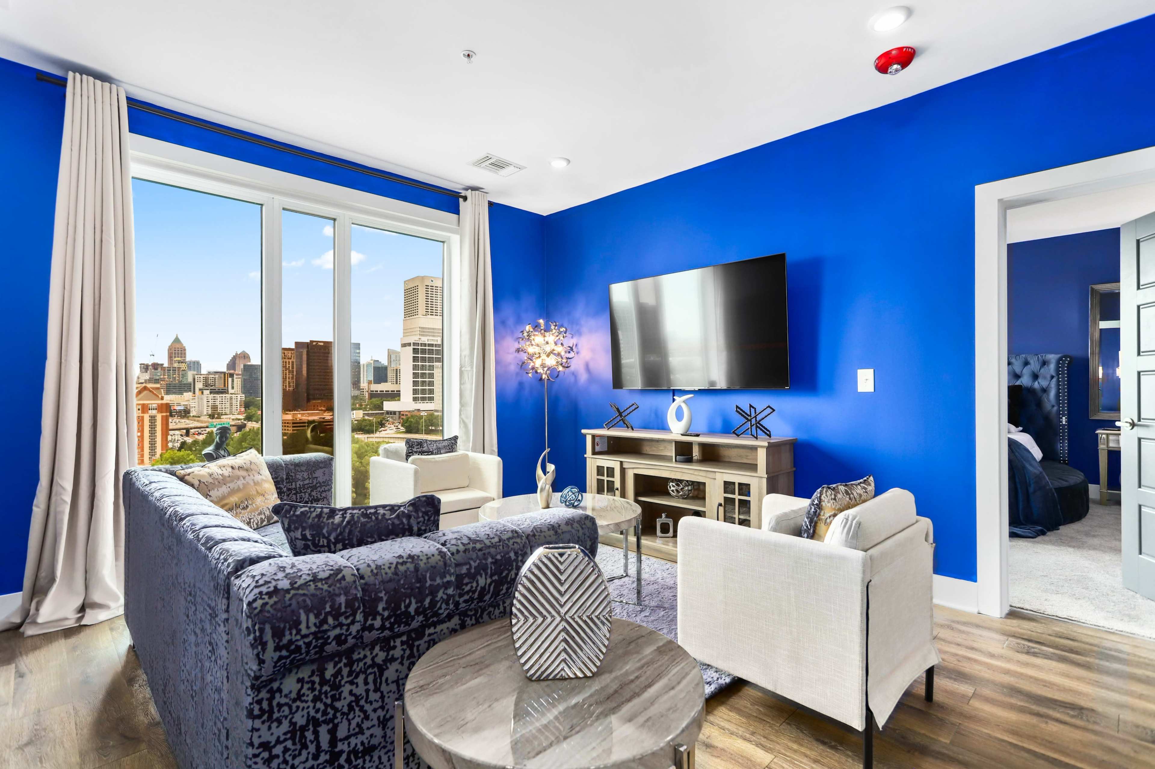 A modern living room features blue walls, a large window with city views, and contemporary furniture including a stylish sofa, armchairs, and a television.