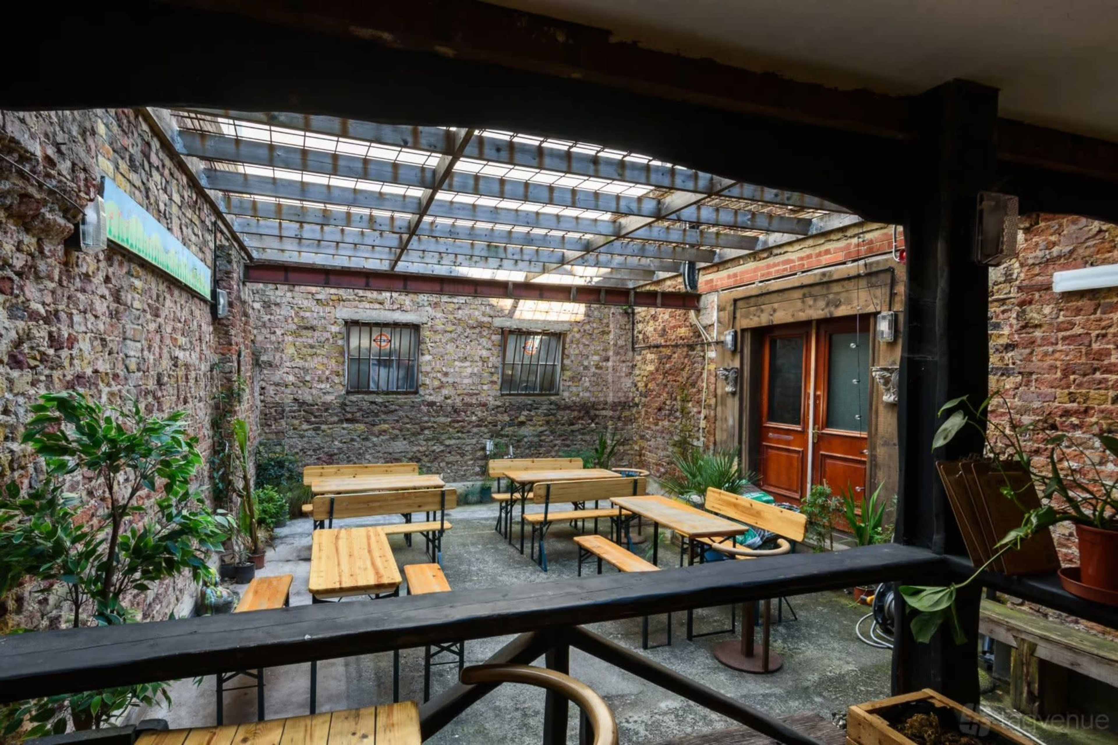 The image shows an empty courtyard with wooden tables and chairs arranged beneath a glass roof, surrounded by brick walls and greenery.