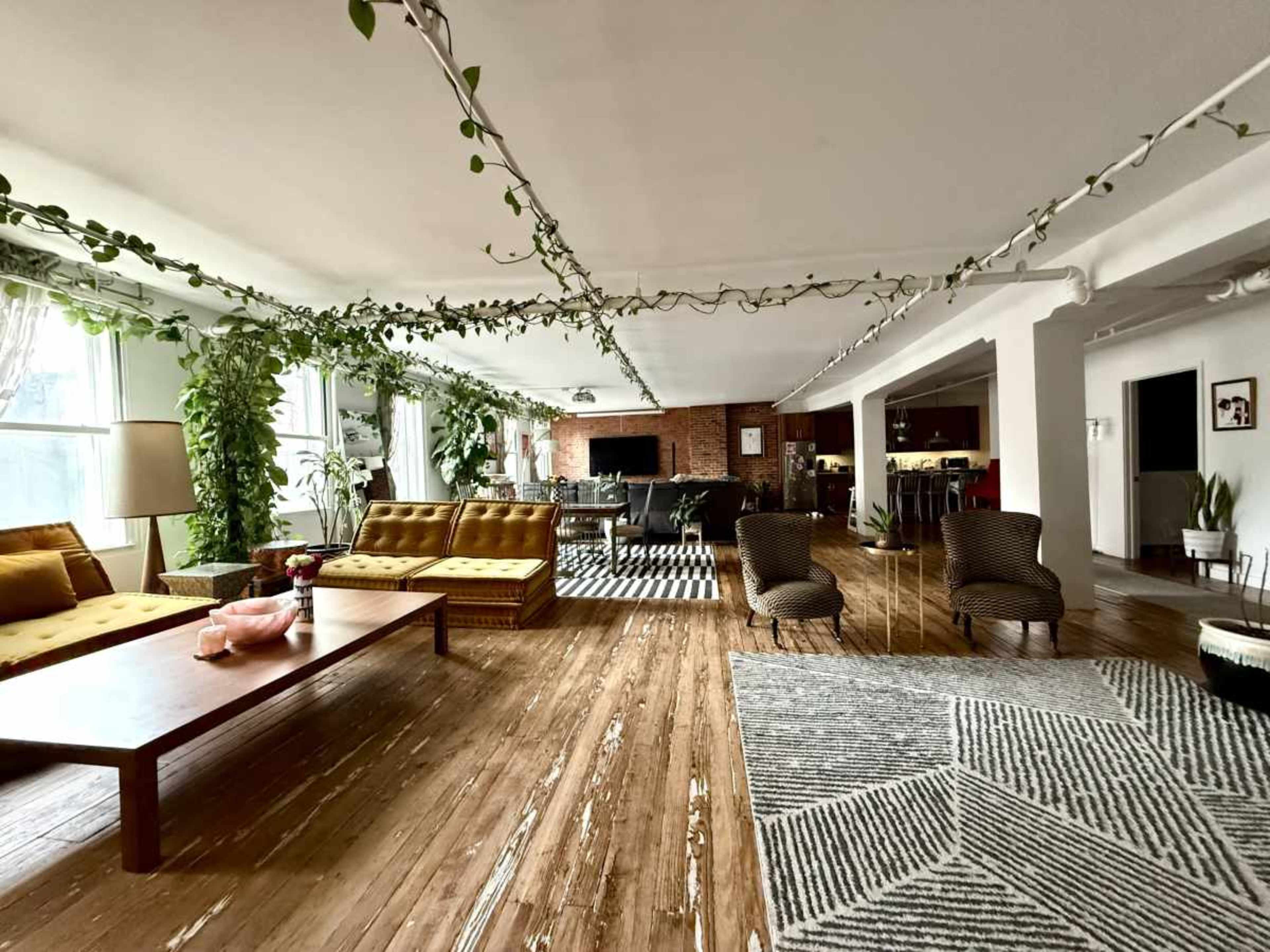 The image shows a spacious living area with wooden floors, large windows, and several plants, featuring two yellow couches, a wooden coffee table, and a patterned rug.