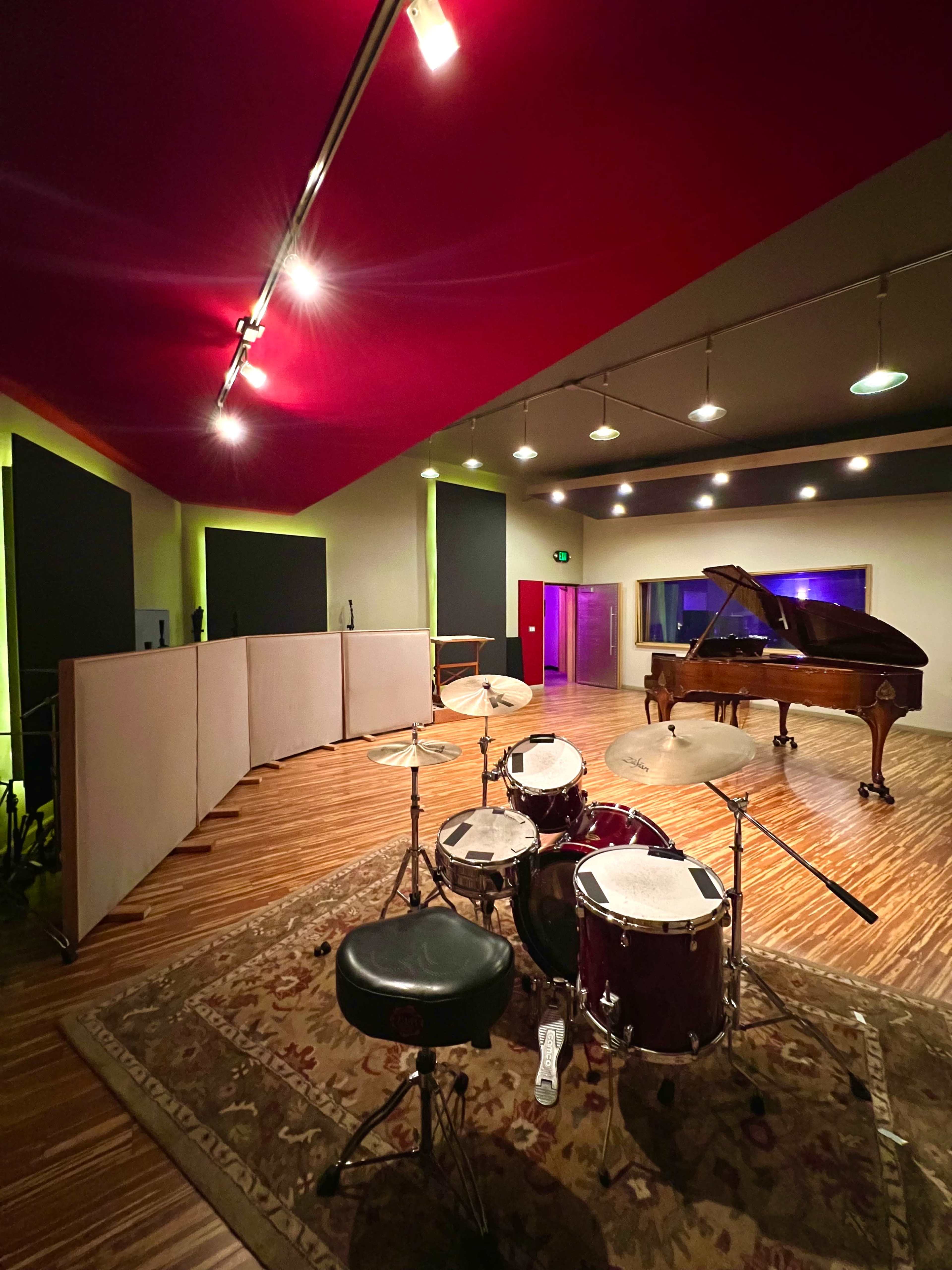 The image shows a music studio featuring a drum set in the foreground and a grand piano positioned against a wall, with sound panels installed around the room.