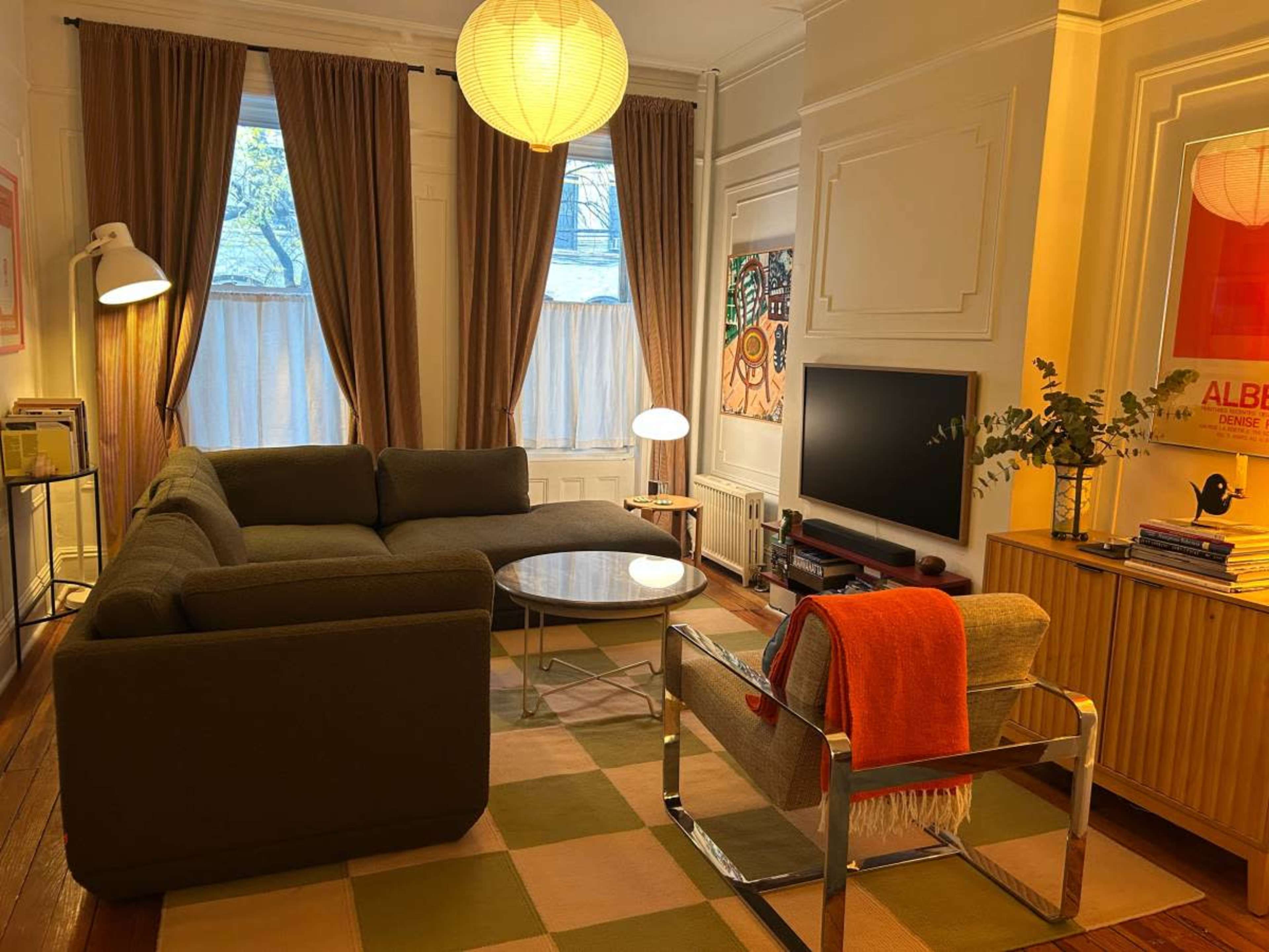 A cozy living room features a sectional sofa, a circular coffee table, a television, and decorative curtains, illuminated by a pendant lamp.