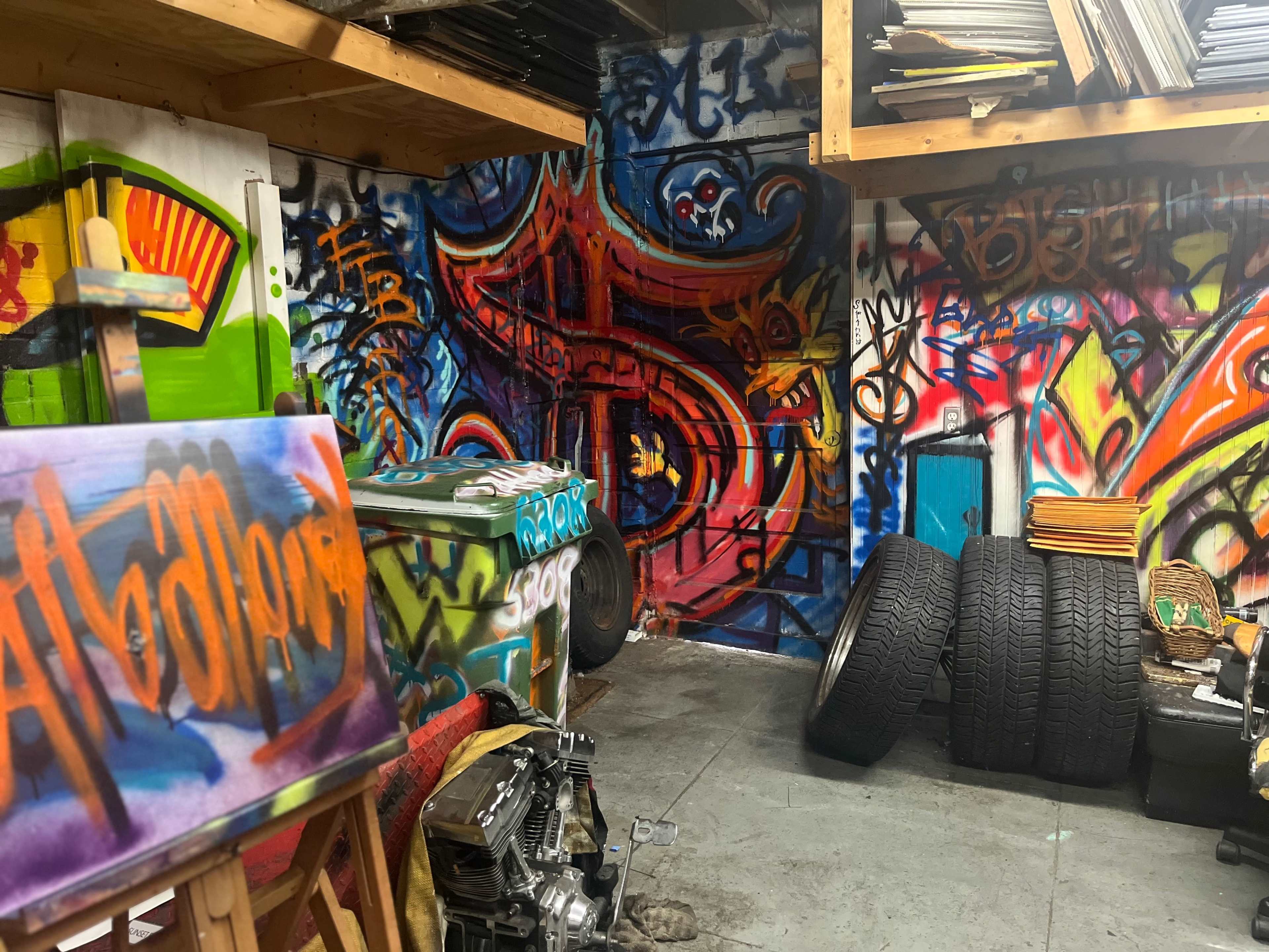 A graffiti-covered workspace featuring a partially visible vehicle, tires, and an easel with a colorful painting.