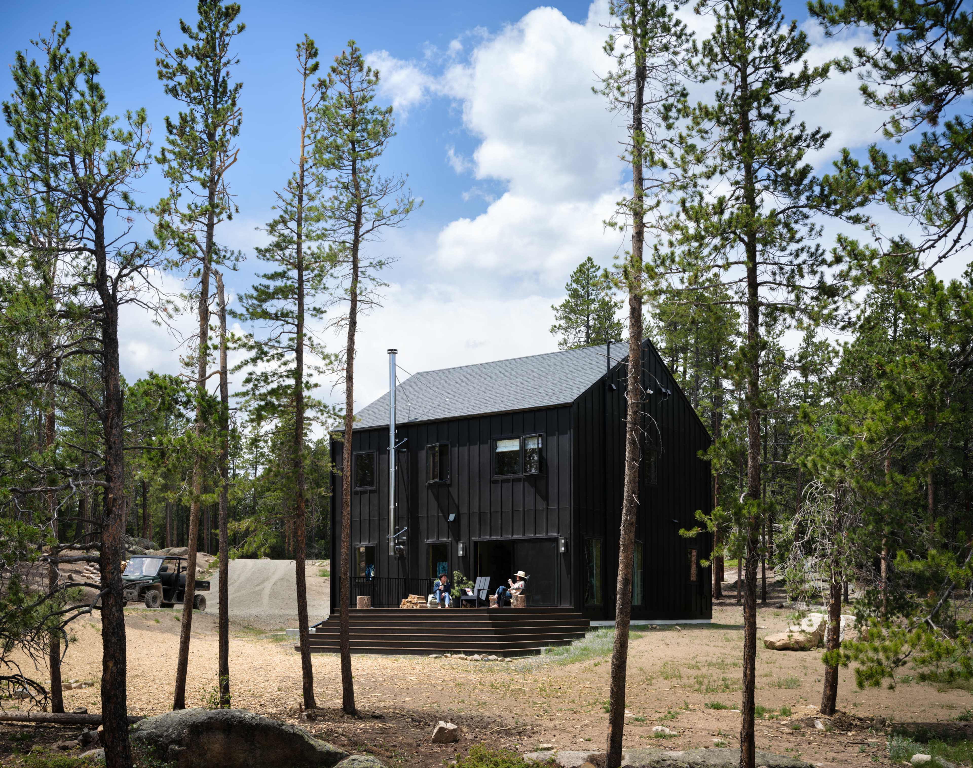 A modern black house stands among tall pine trees, with a small gathering on the front deck and a vehicle parked nearby.