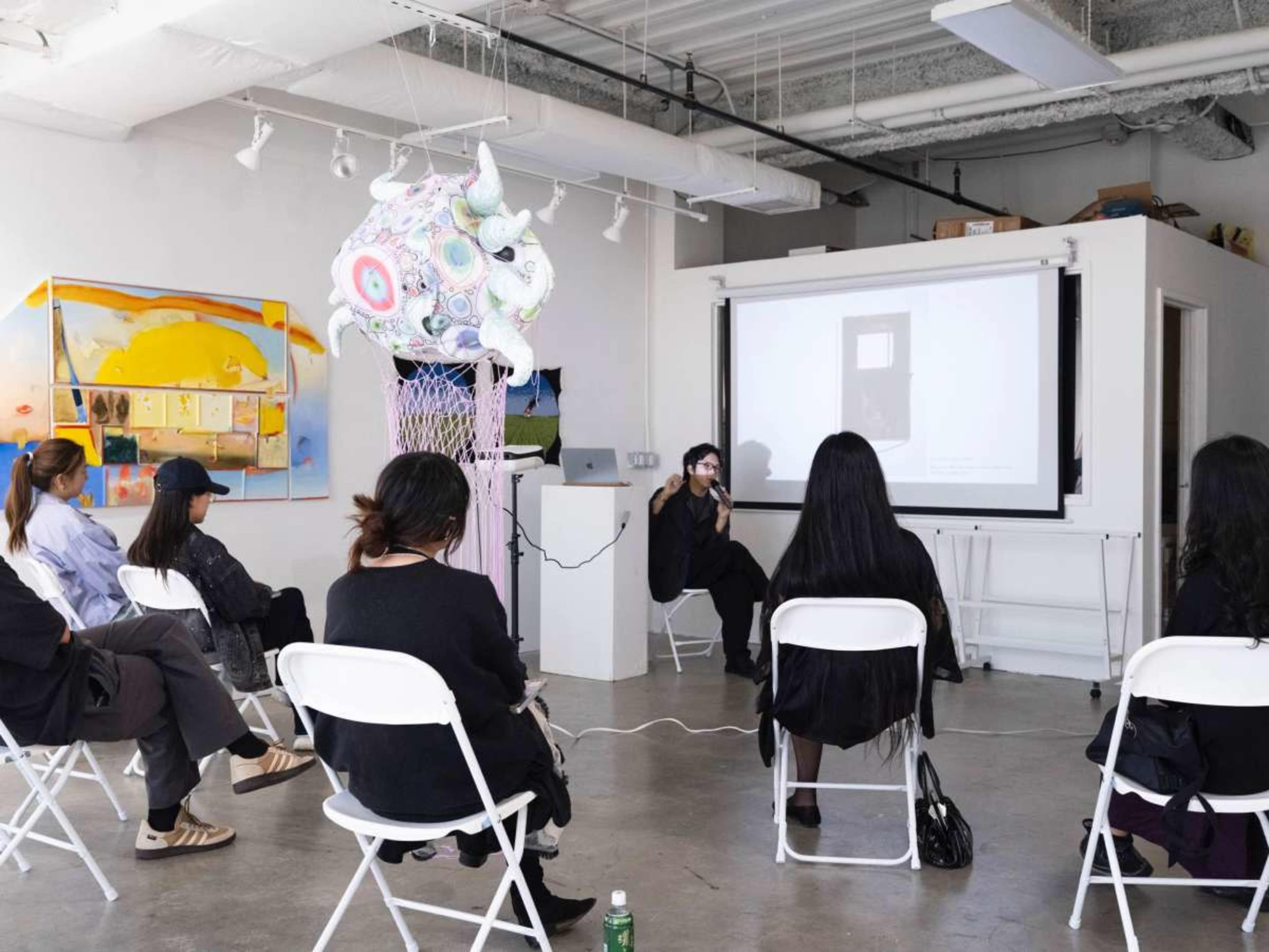 A group of people sits on white chairs in a gallery-like space while a speaker presents on a screen at the front.