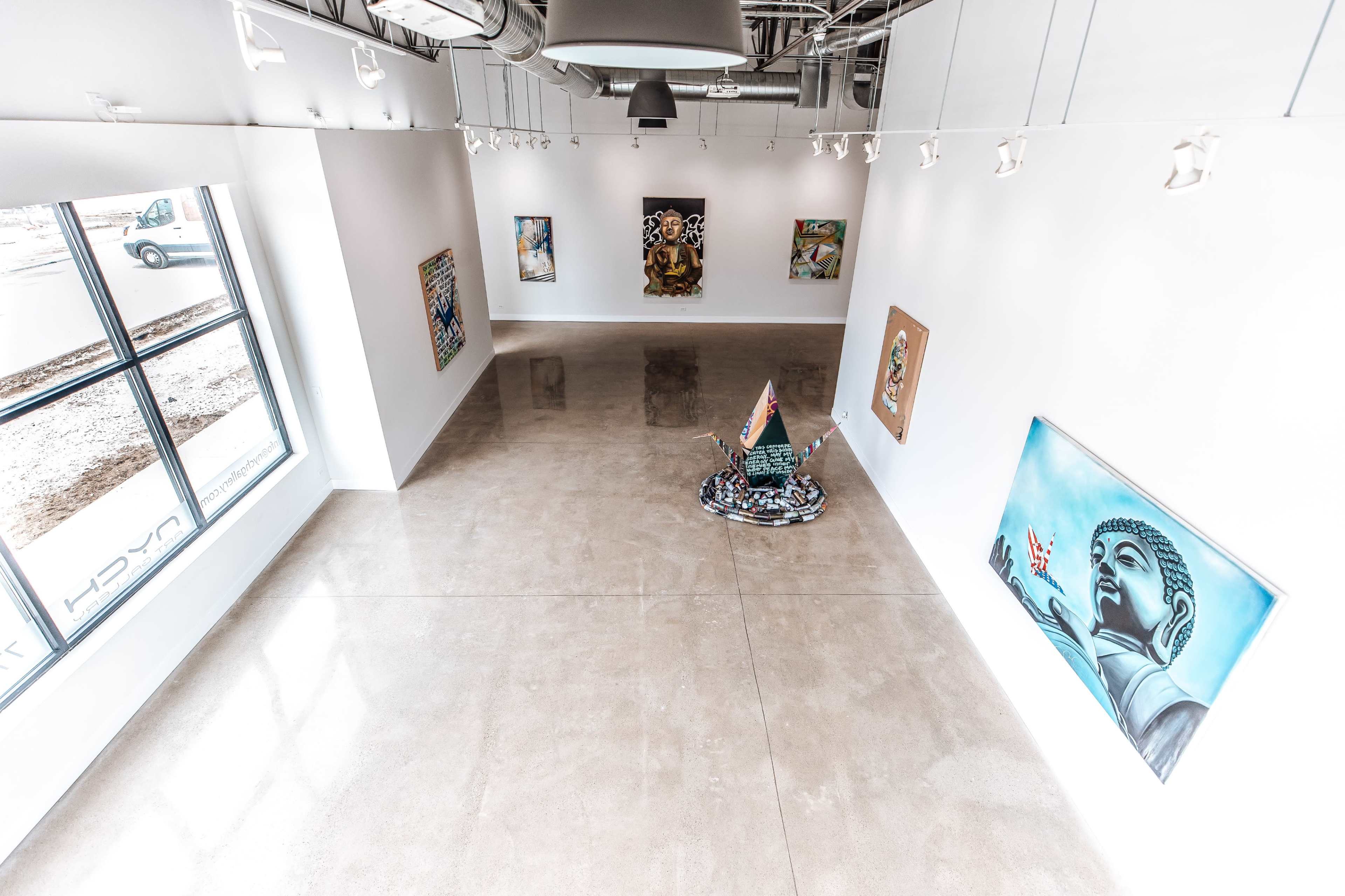 The image shows an art gallery with polished concrete floors, featuring several framed artworks on white walls and a central installation.