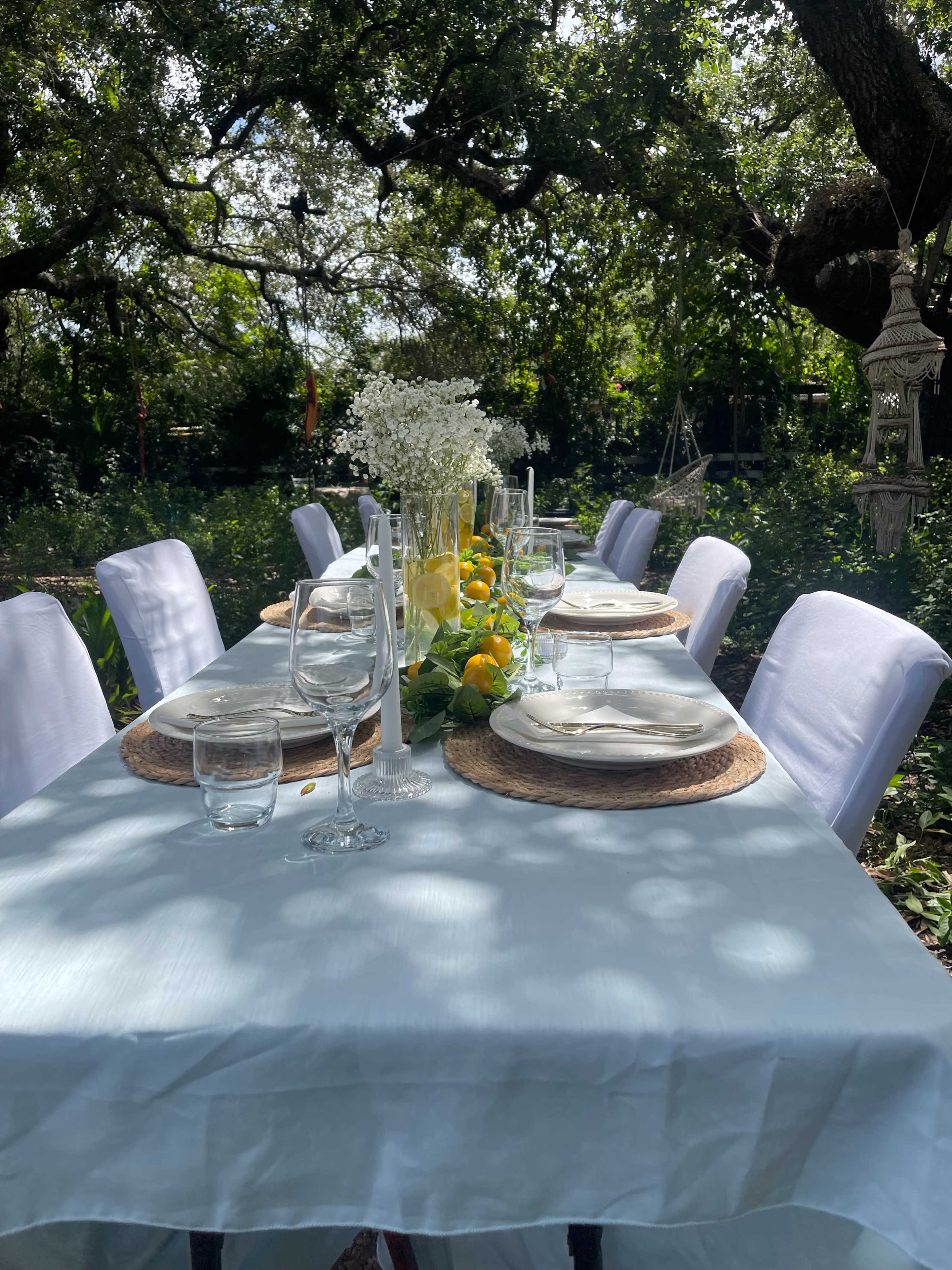 A long table is set outdoors with white tablecloths, glassware, and decorative centerpieces of flowers and lemons under the shade of trees.
