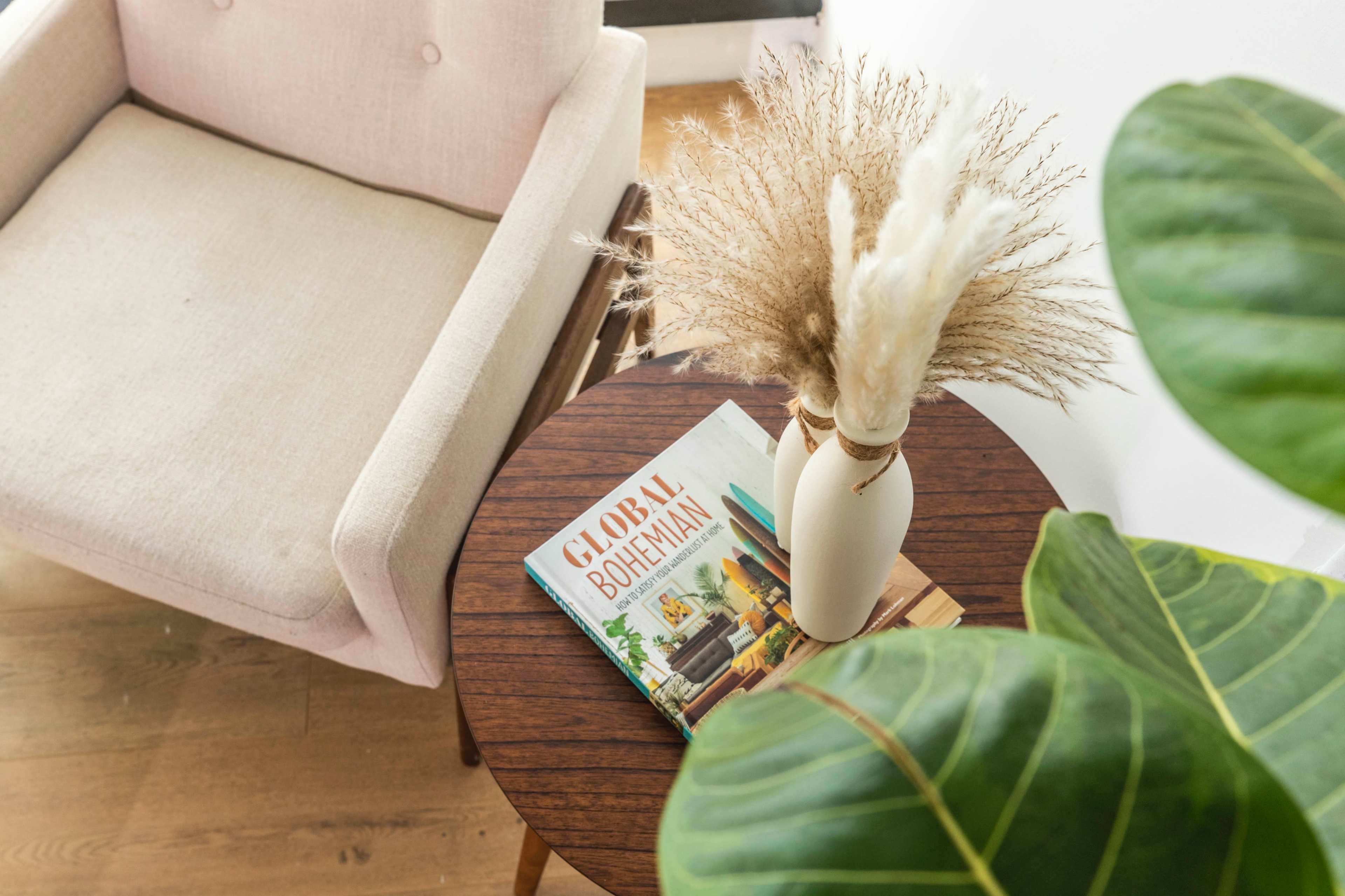A light-colored armchair is positioned next to a wooden side table, which holds a magazine titled "Global Bohemian" and a vase with dried plants, while a large green leaf partially obscures the view.