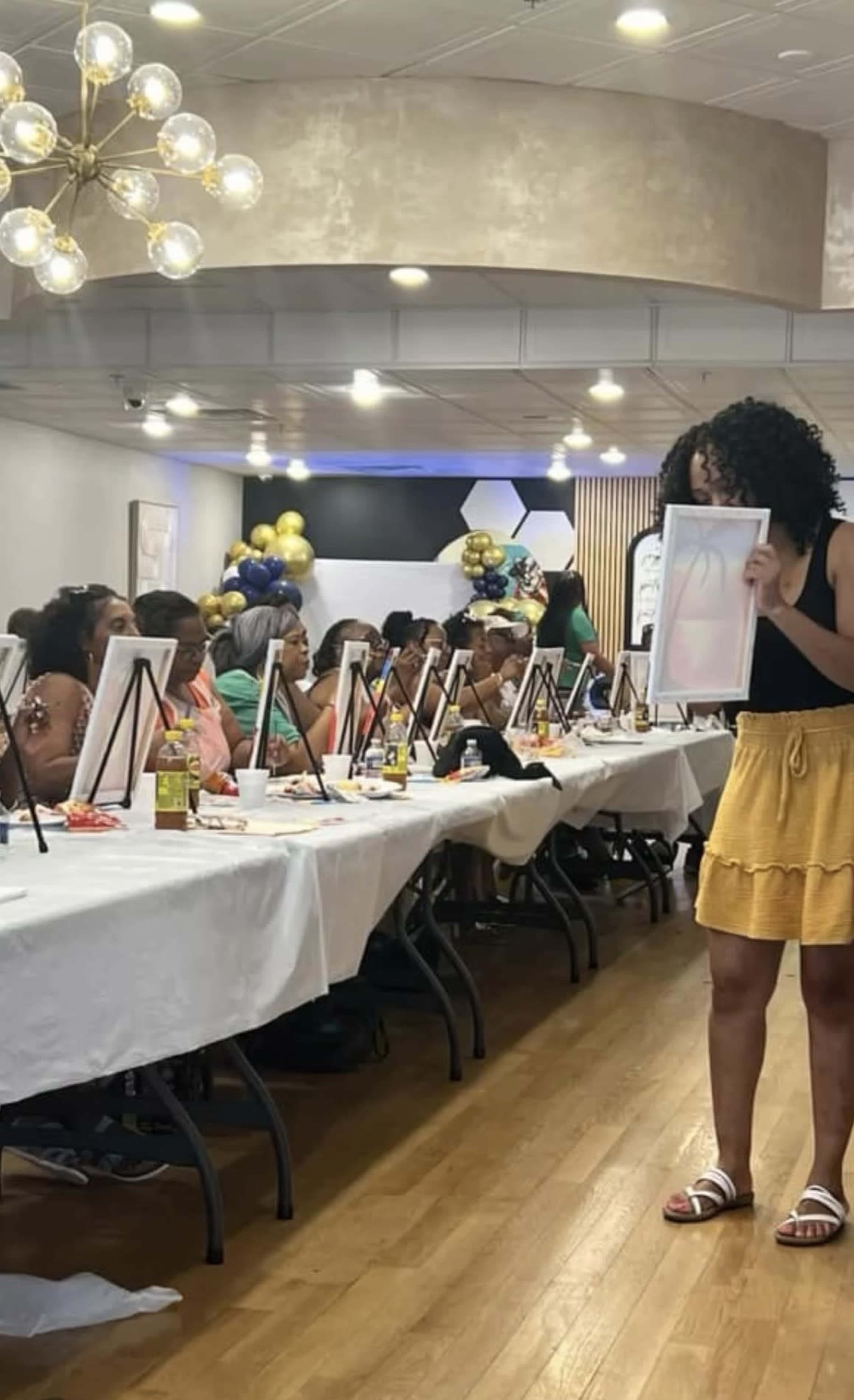 A woman stands at the front of a banquet hall holding up a piece of artwork, while guests seated at long tables look on in an event setting.