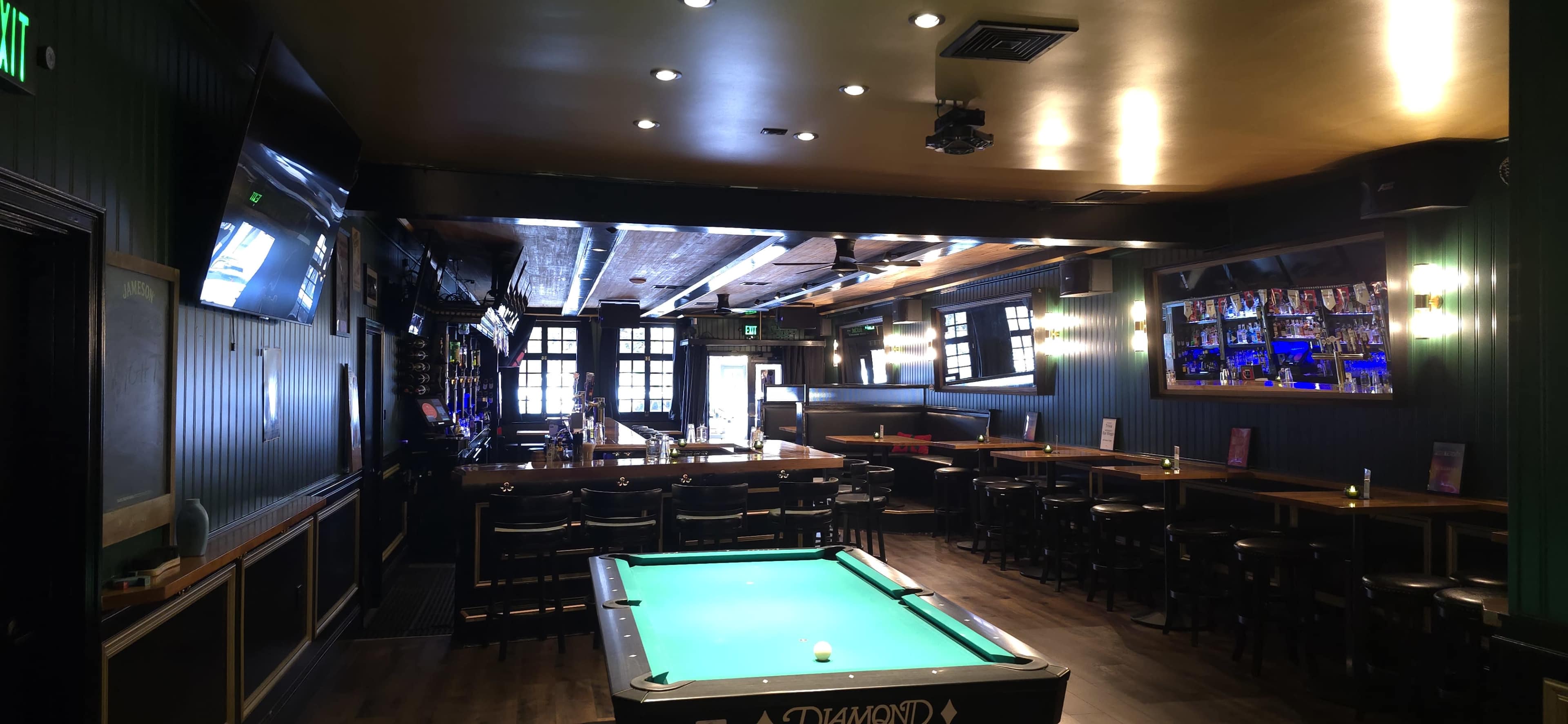 The image shows a spacious bar with a green pool table, wooden tables and chairs, and a long bar counter, all under warm overhead lighting.