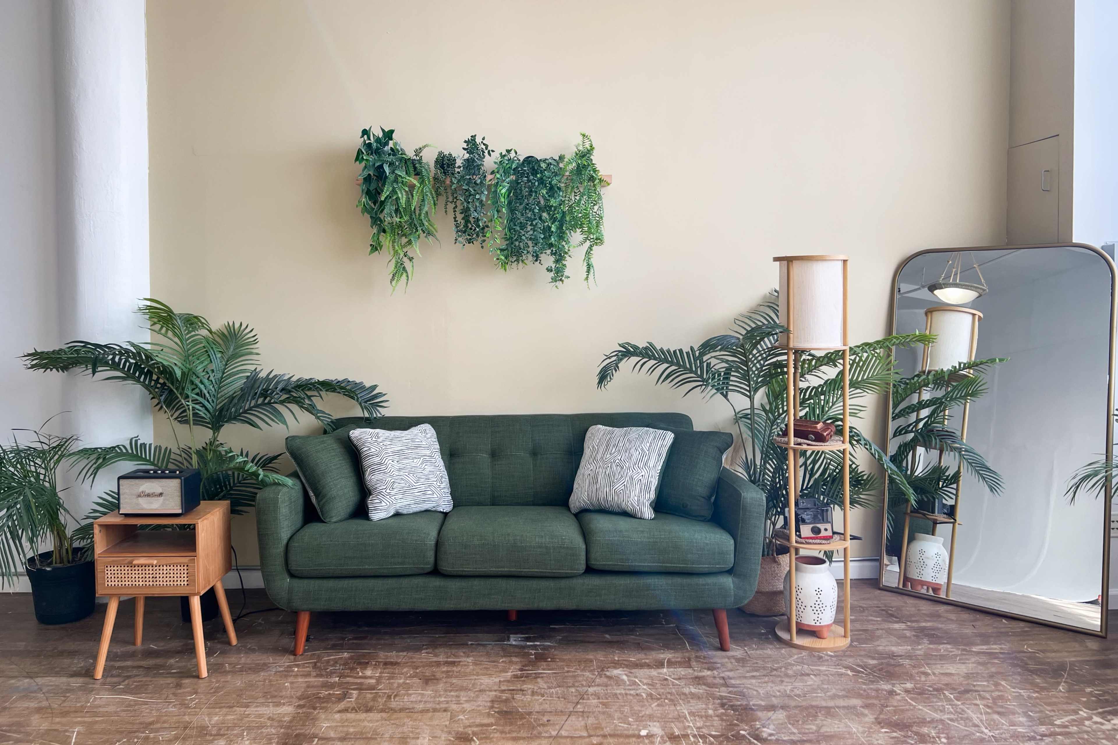A green sofa sits against a light-colored wall, accompanied by potted plants, a wooden side table, and a large mirror.