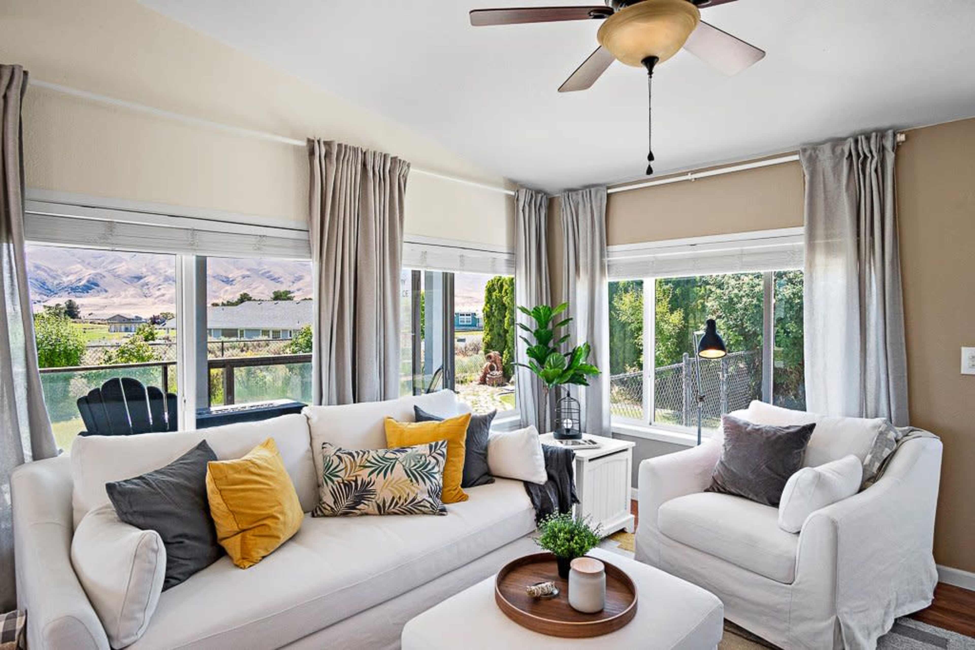 A bright living room features a white sectional sofa with decorative pillows, large windows offering a view of a landscape, and a round coffee table in the center.