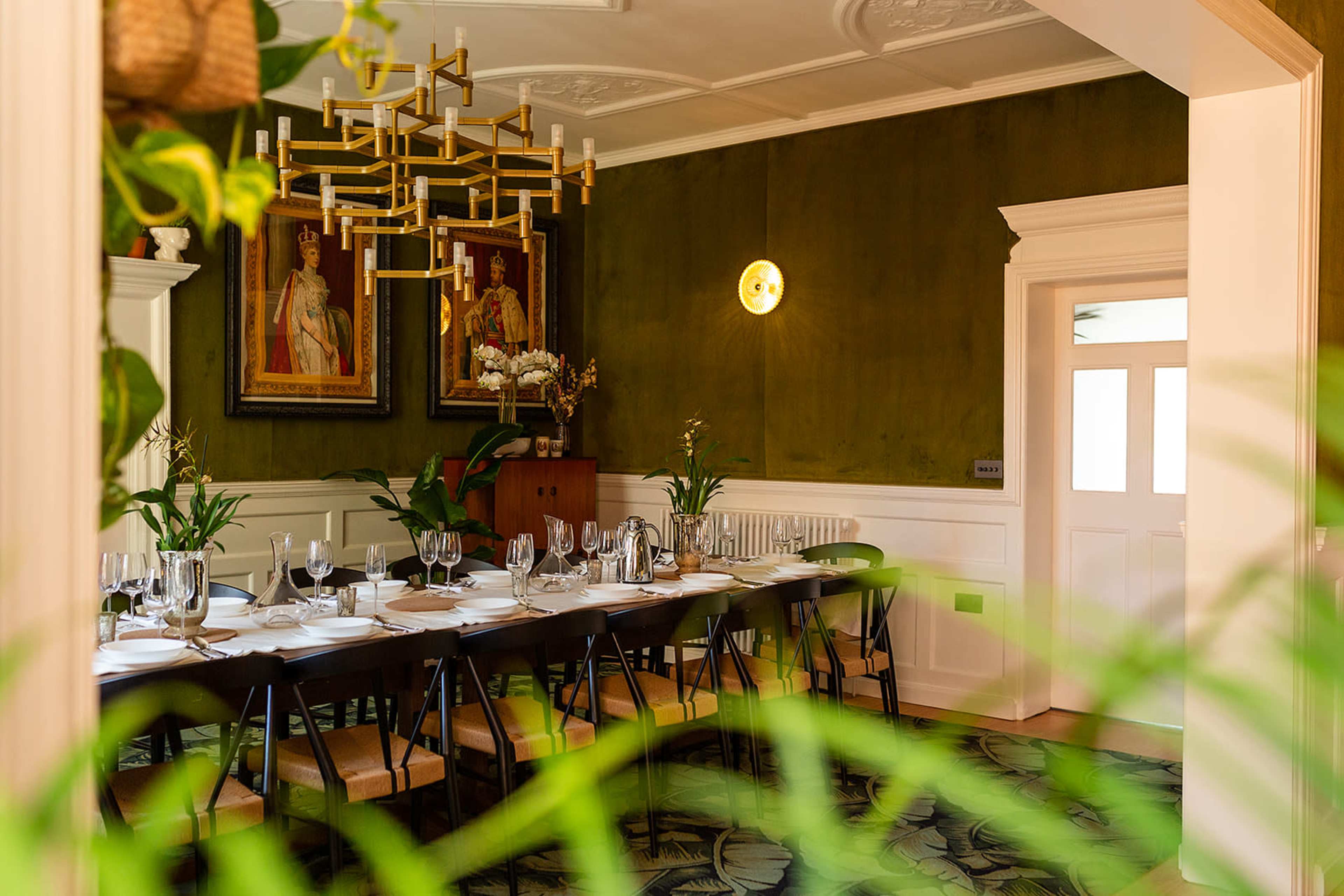 A long dining table is set with tableware in a room featuring green walls, chandeliers, and framed portraits.