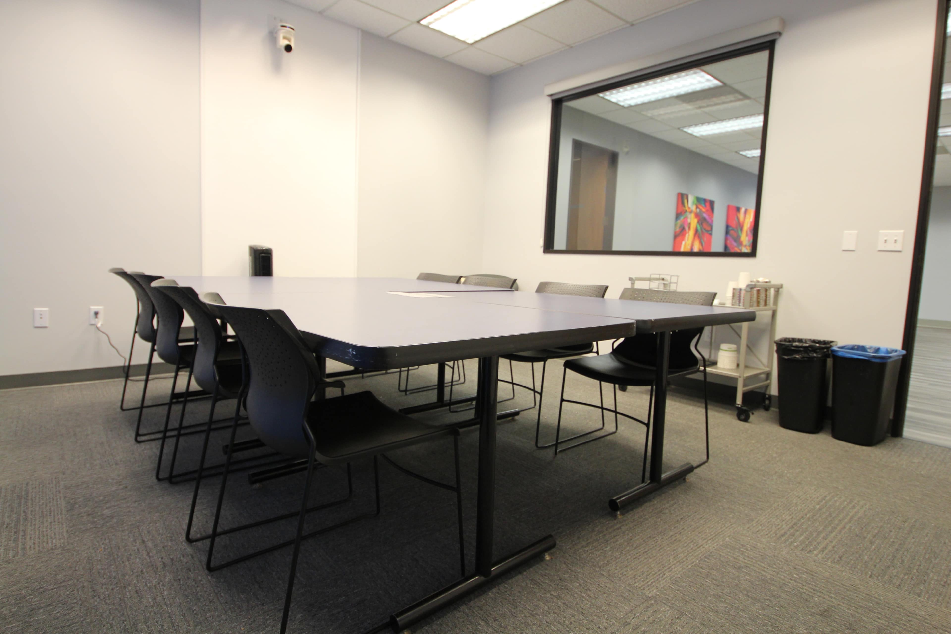 A conference room features a large table surrounded by black chairs, with a window overlooking an abstract painting and a garbage bin in the corner.