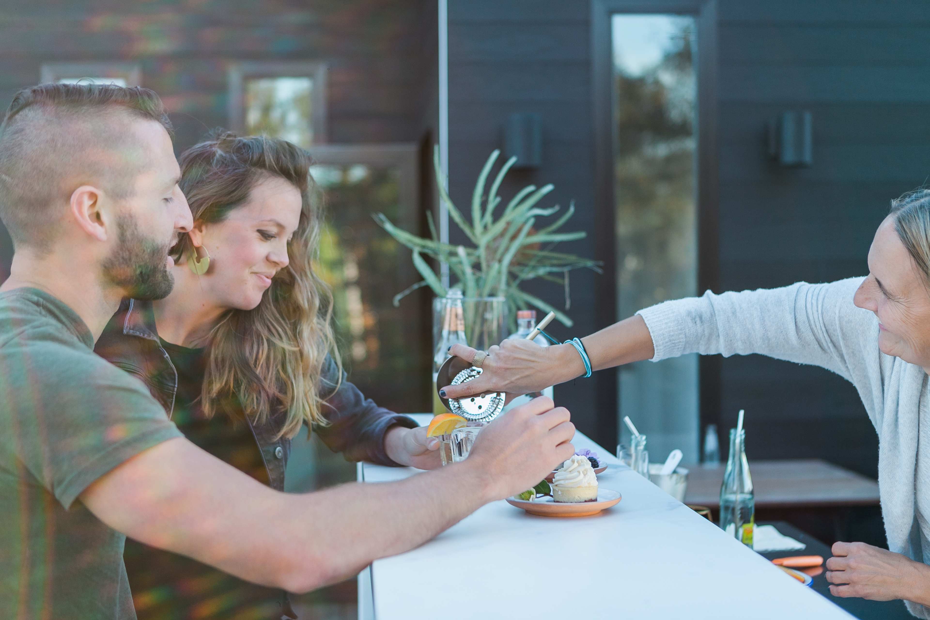 Two people are sitting at a counter while a third person serves them drinks.