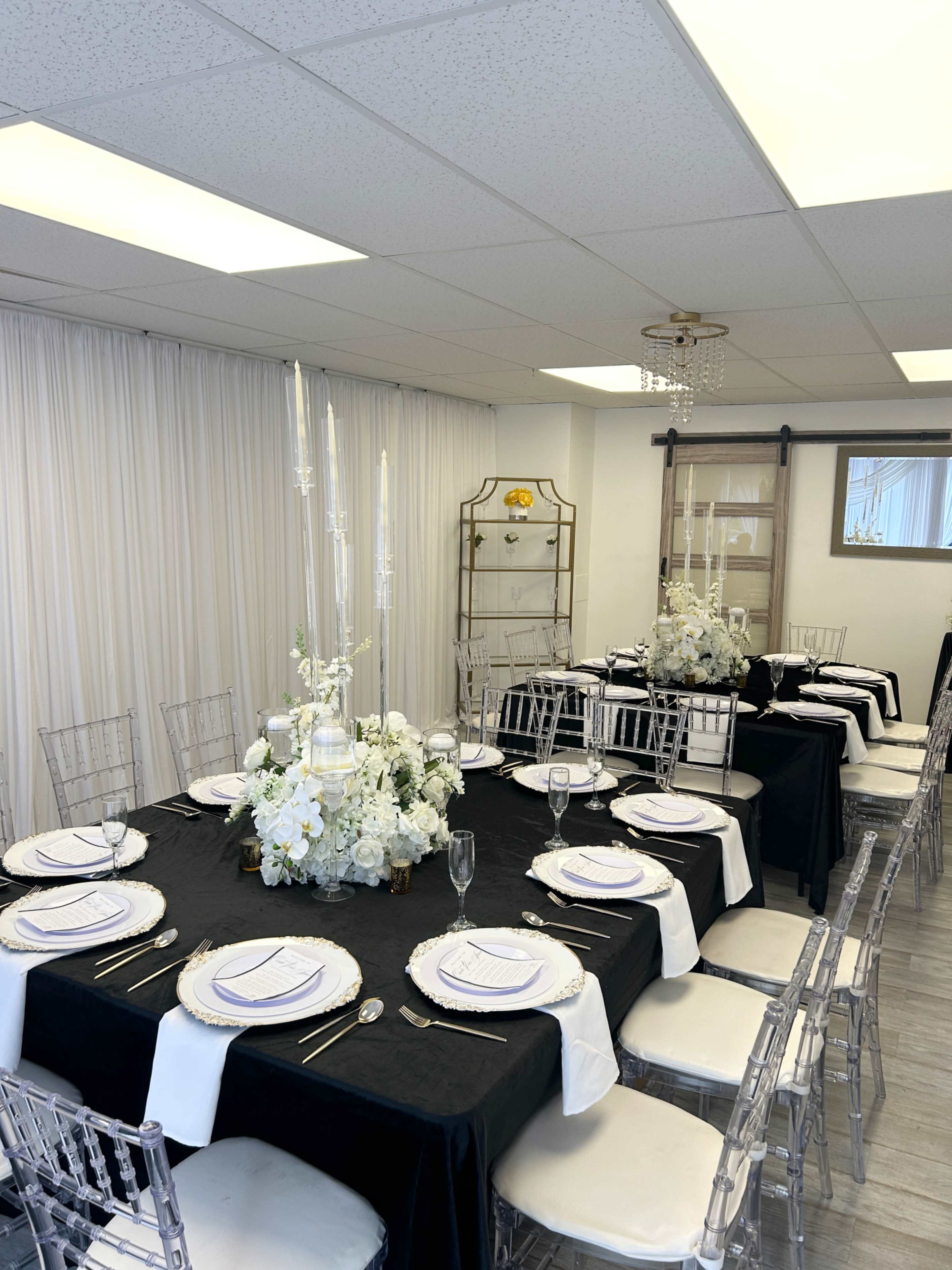 A long dining table is elegantly set with black and white tablecloths, floral centerpieces, and clear chiavari chairs in a well-lit room.