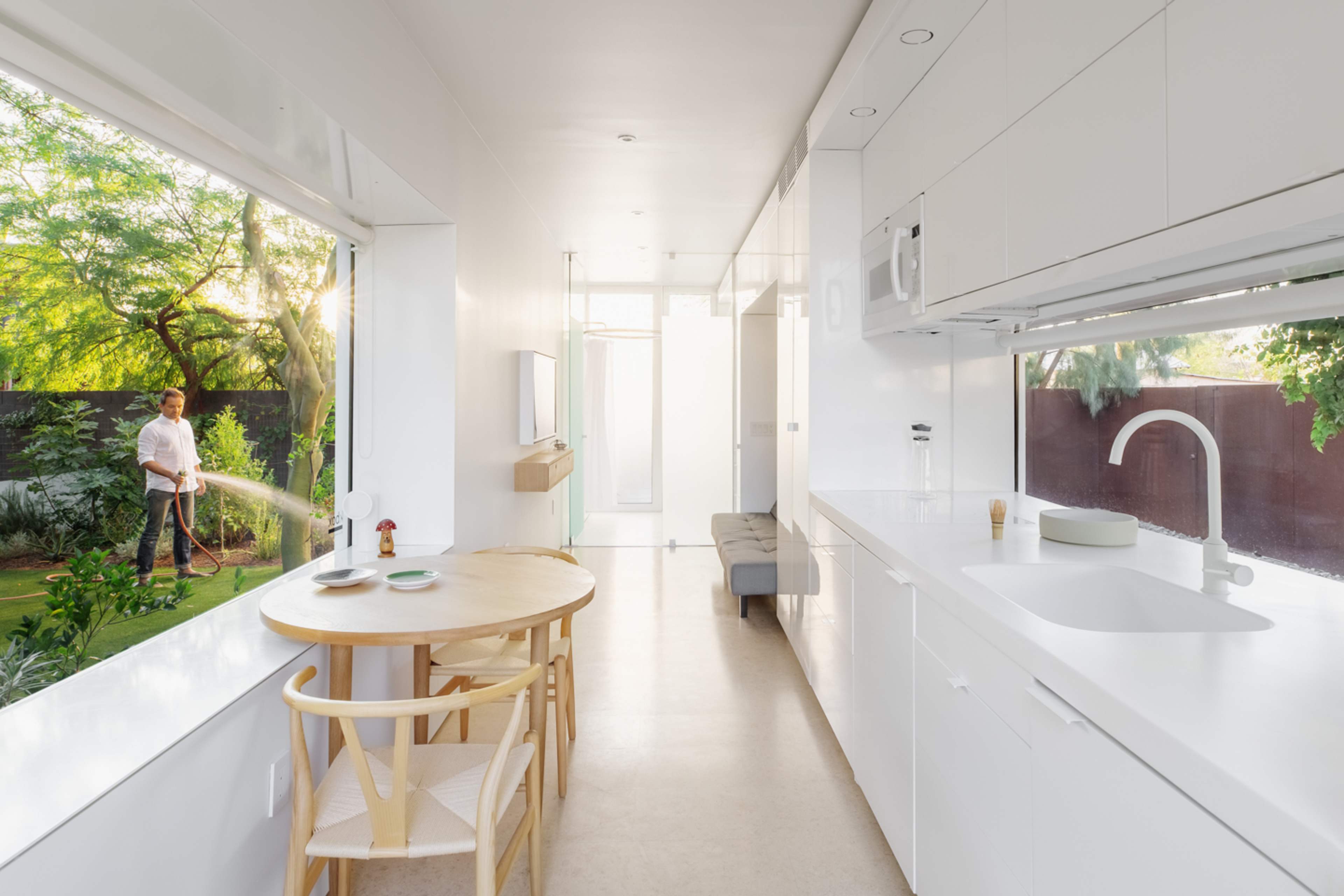 A bright, modern kitchen features a dining table and a view of a person watering plants in a lush garden outside.