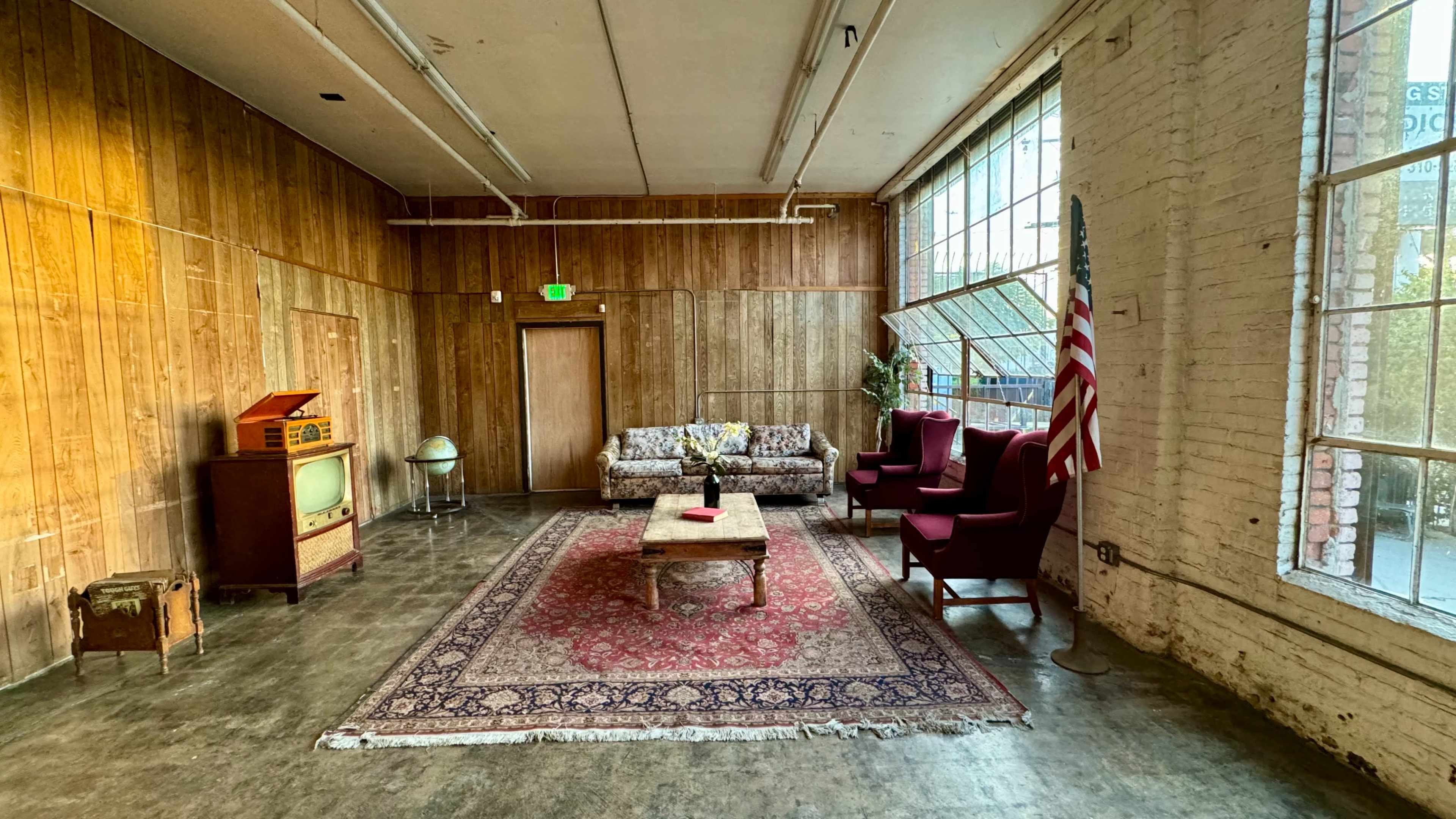 A cozy room features a patterned rug, a wooden wall, a vintage radio, a green globe, and an American flag next to a couch and two armchairs.