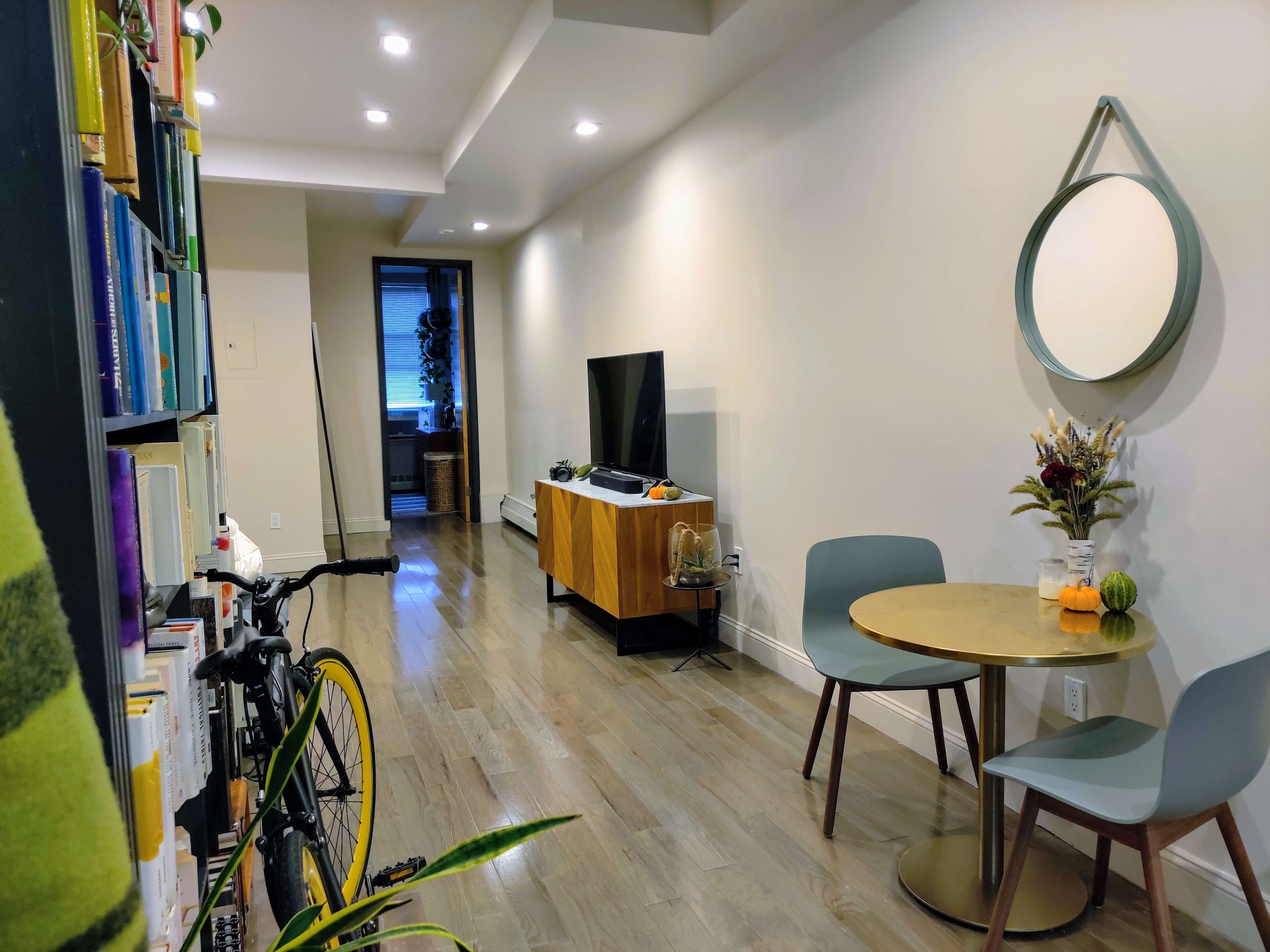 A cozy living space with a bicycle parked beside a bookshelf, a dining table set for two, and a television against a light-colored wall.