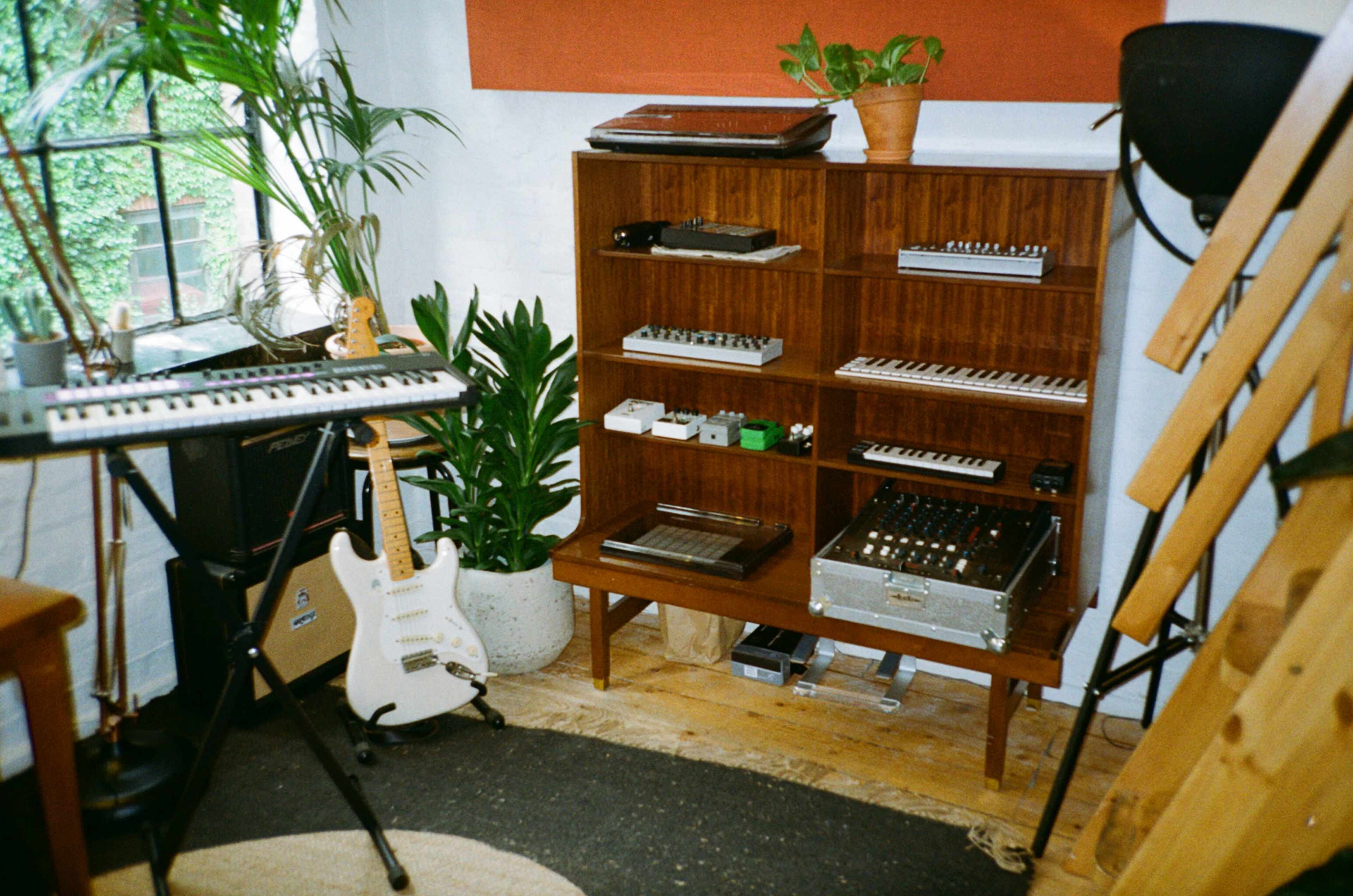 A cozy music studio featuring a wooden shelving unit filled with electronic instruments, a white guitar leaning against the wall, and various plants.