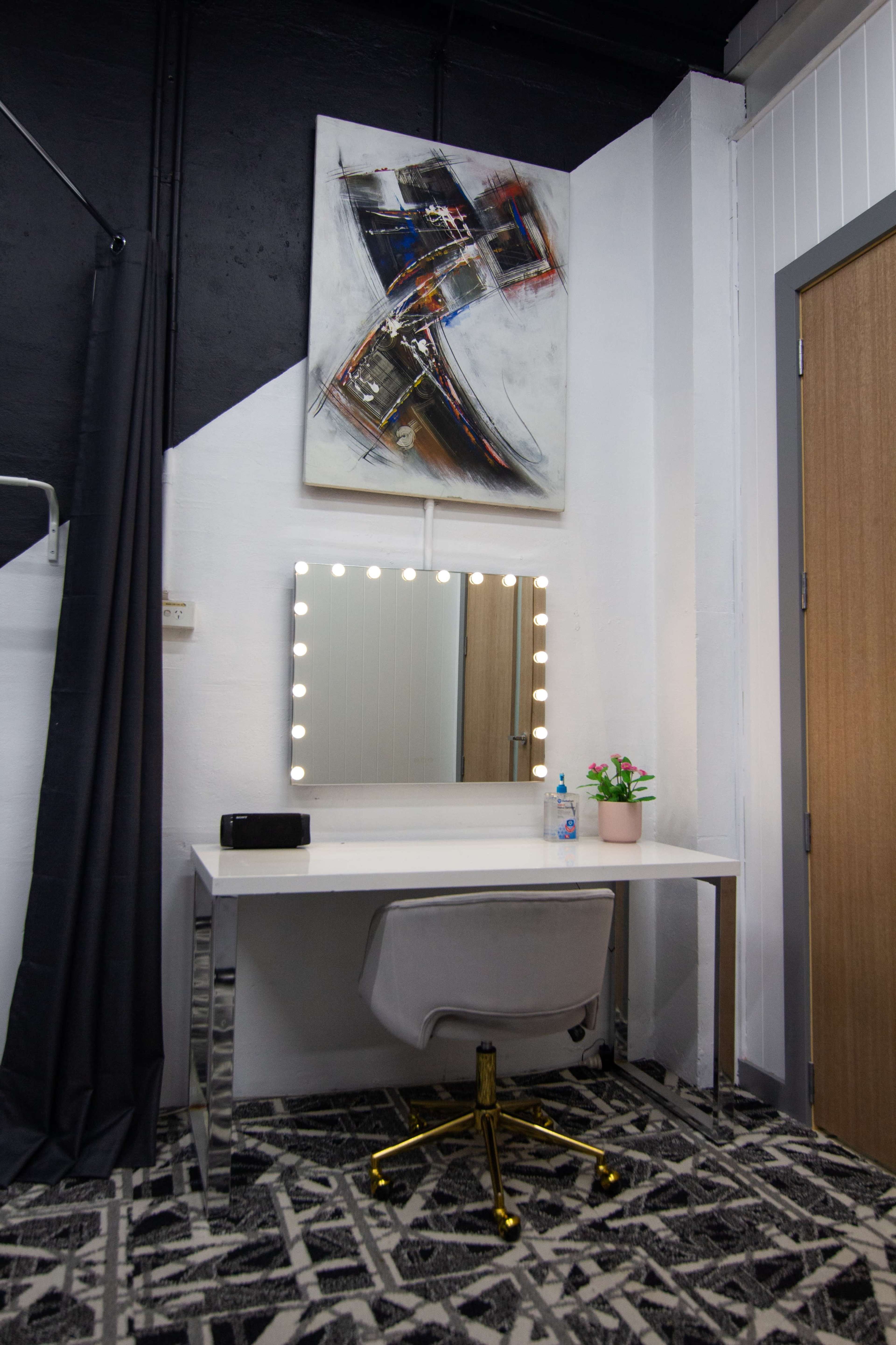 A modern vanity desk with a large mirror surrounded by lights, a small plant, and a chair, set against a dark wall with an abstract painting above.
