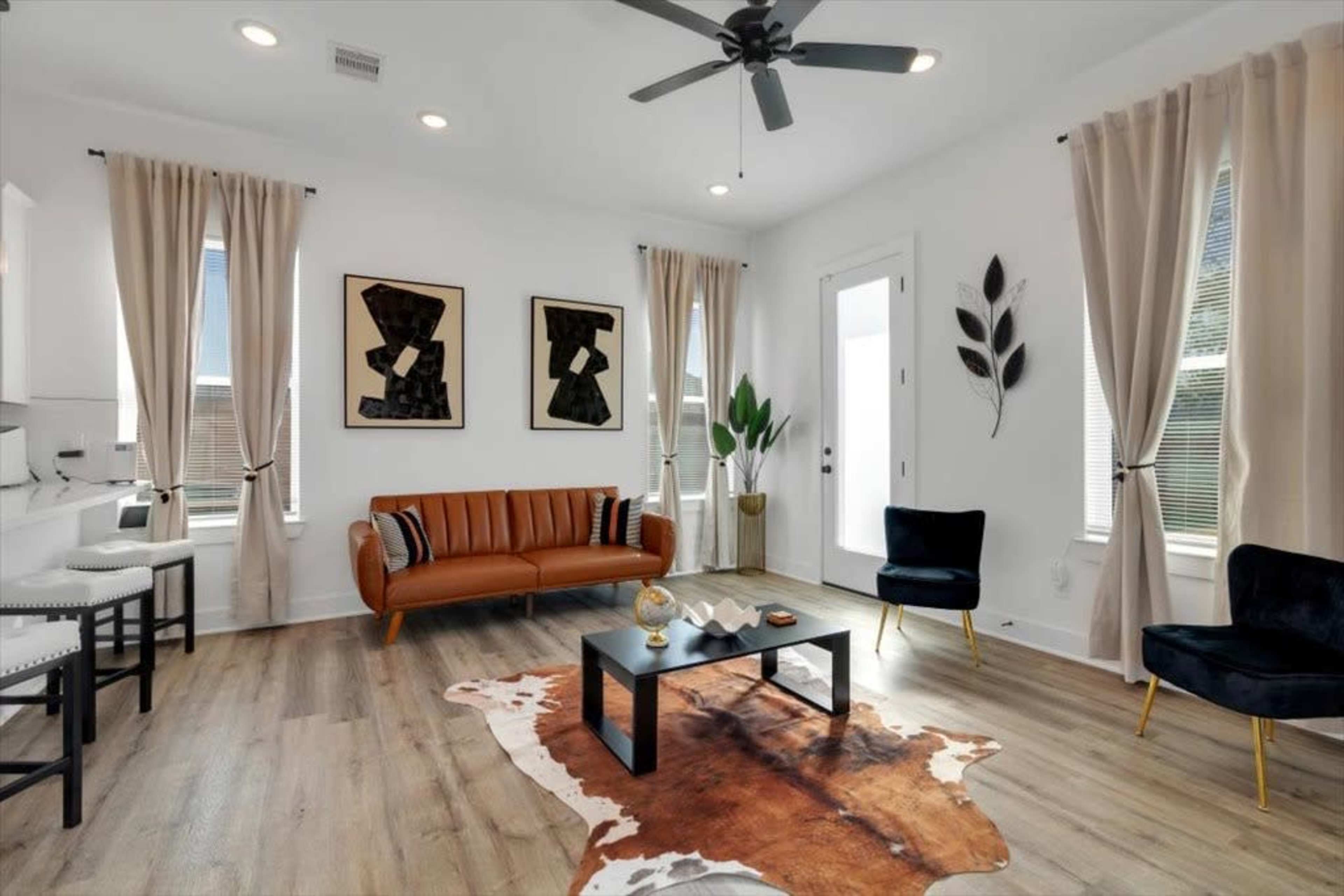 The living room features a brown leather sofa, two black chairs, a wooden coffee table on a cowhide rug, and framed artworks on the white walls.