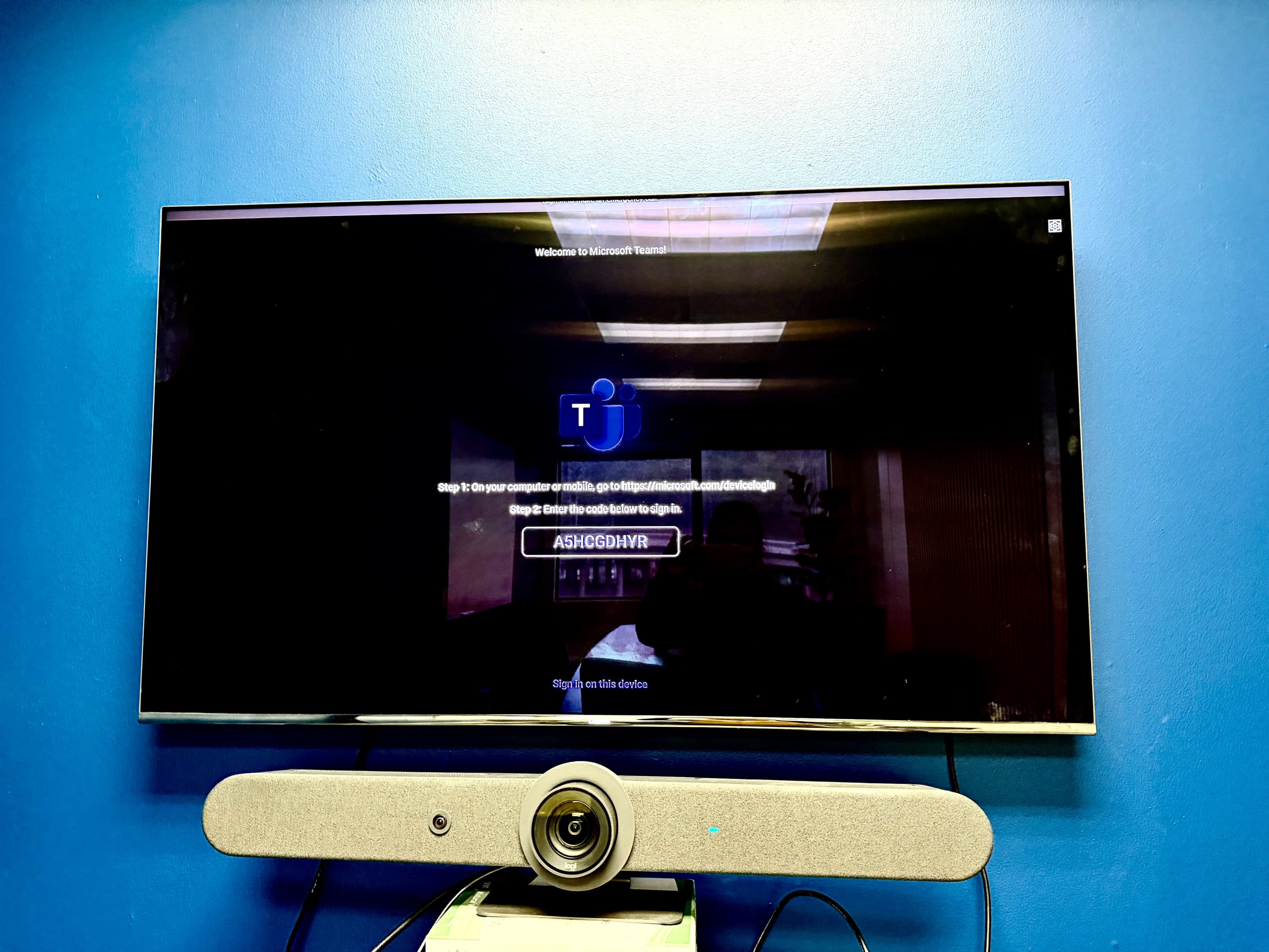 The image shows a television mounted on a blue wall displaying a welcome screen for Microsoft Teams and prompting the user to log in.