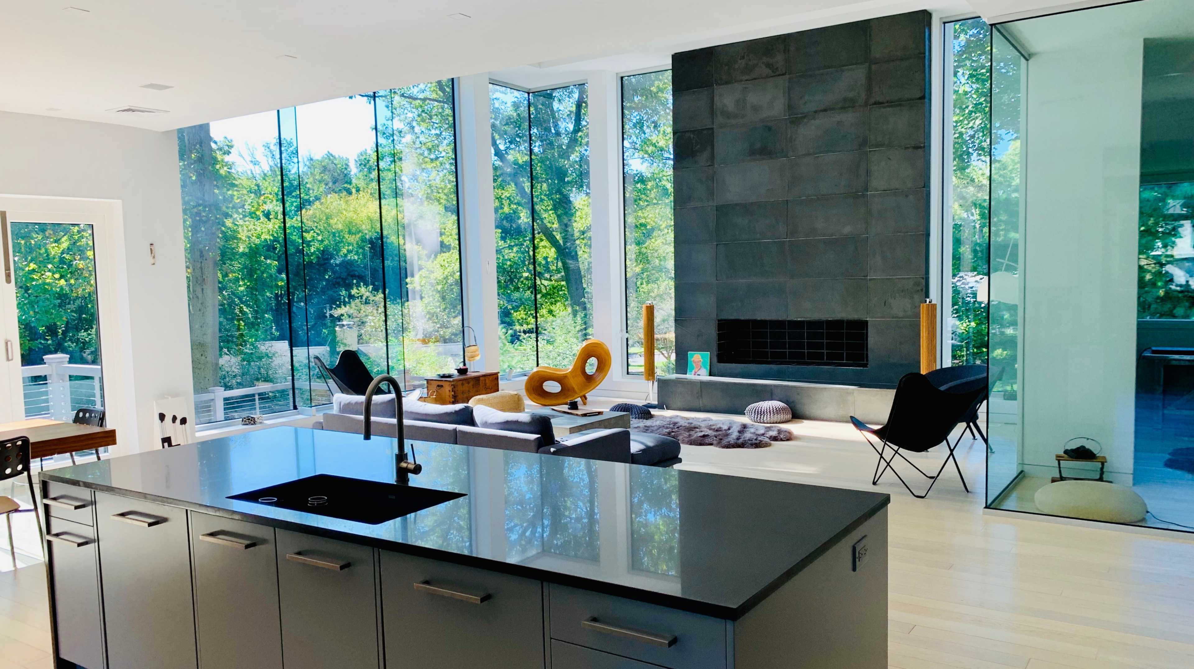 A modern living space features a kitchen with a black countertop, large windows overlooking a wooded area, and a cozy seating arrangement near a stone fireplace.