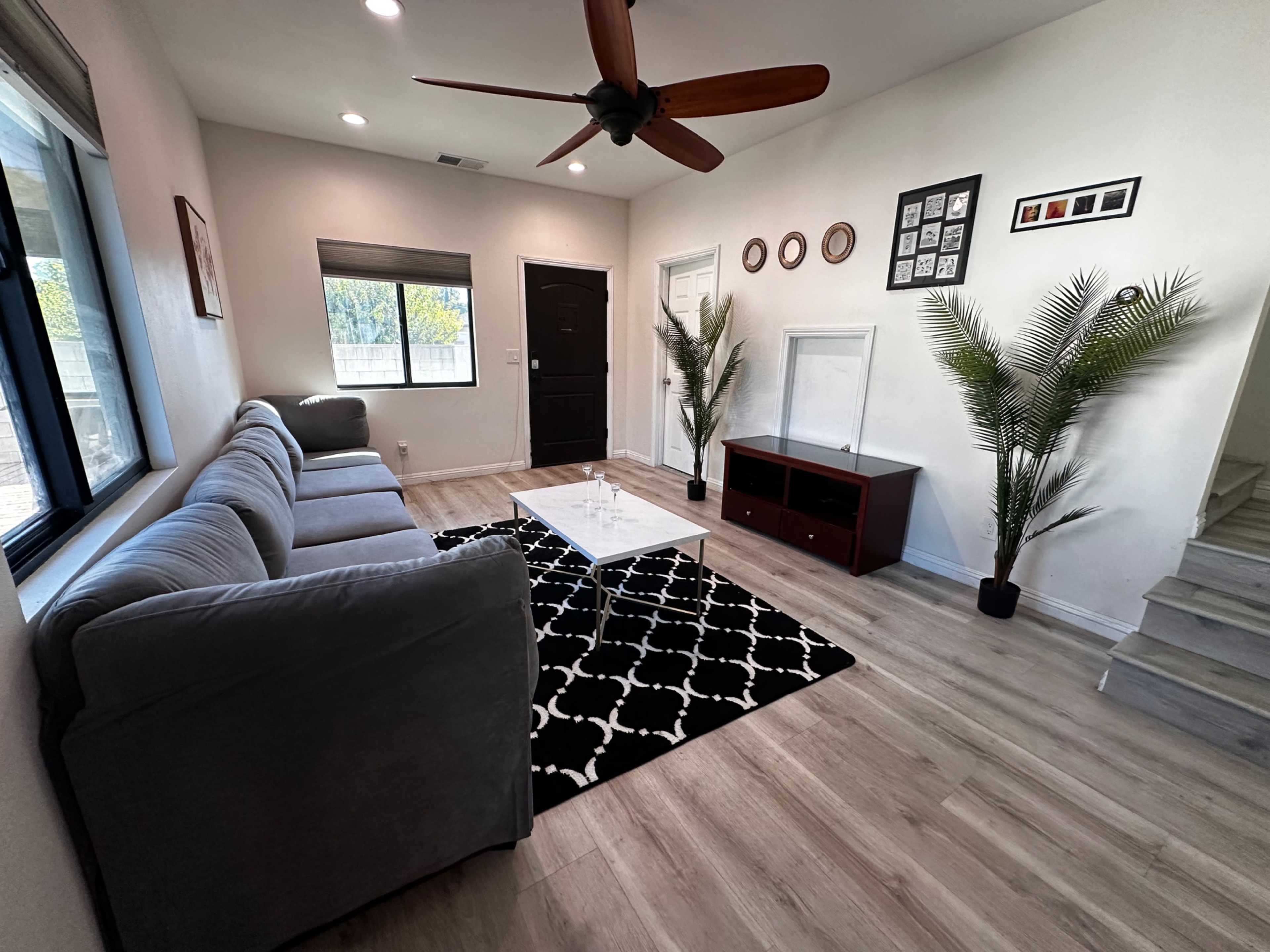 A modern living room with a gray sofa, a glass coffee table, a black and white patterned rug, and decorative plants, alongside a doorway and staircase.