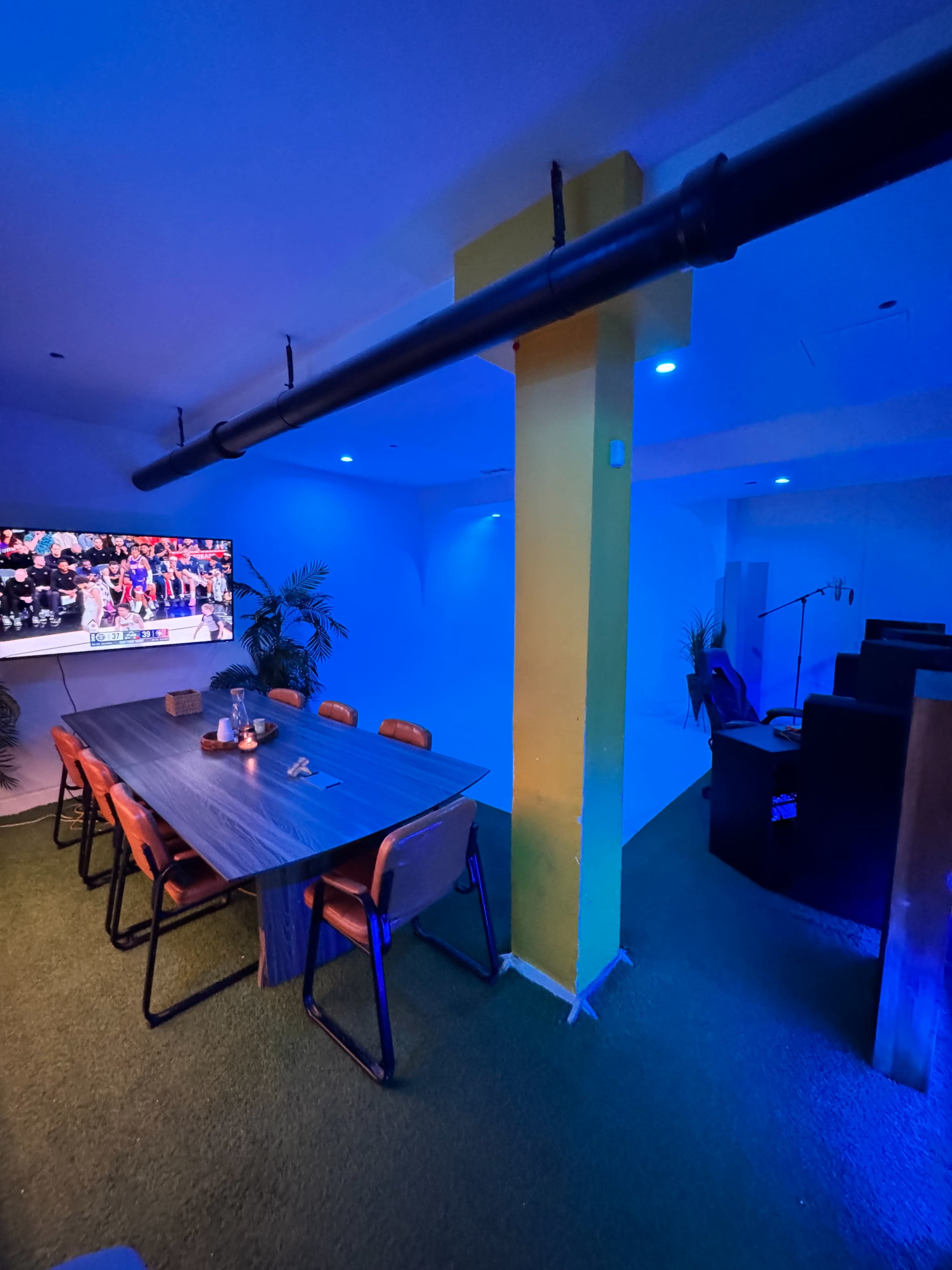 The image shows a modern, brightly lit meeting room featuring a large table with chairs, a screen displaying an event, and decorative plants in a blue and yellow color scheme.