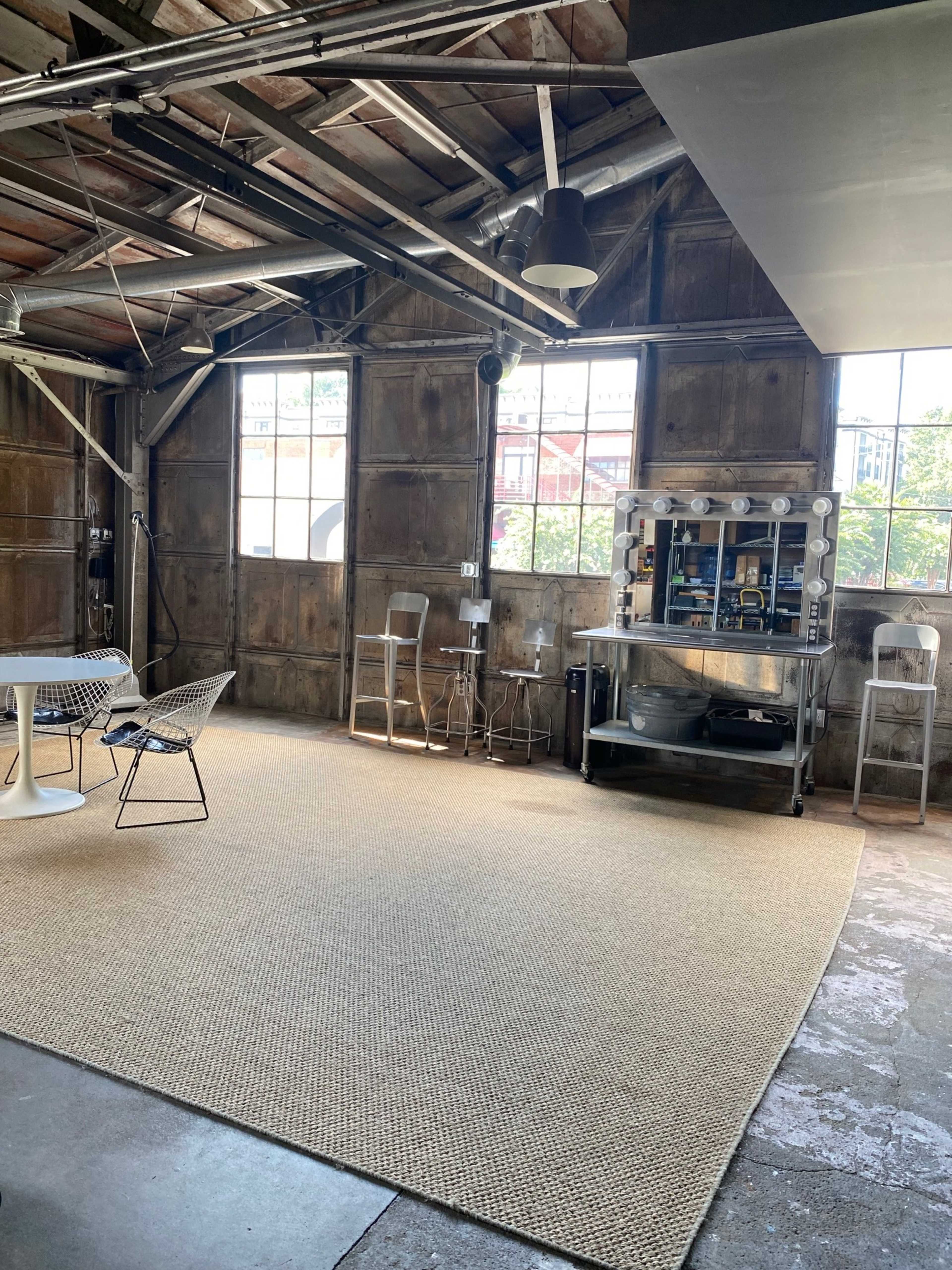 The image shows an industrial-style interior with wooden paneling, large windows, a round table with four chairs, and a metal cart against the wall.