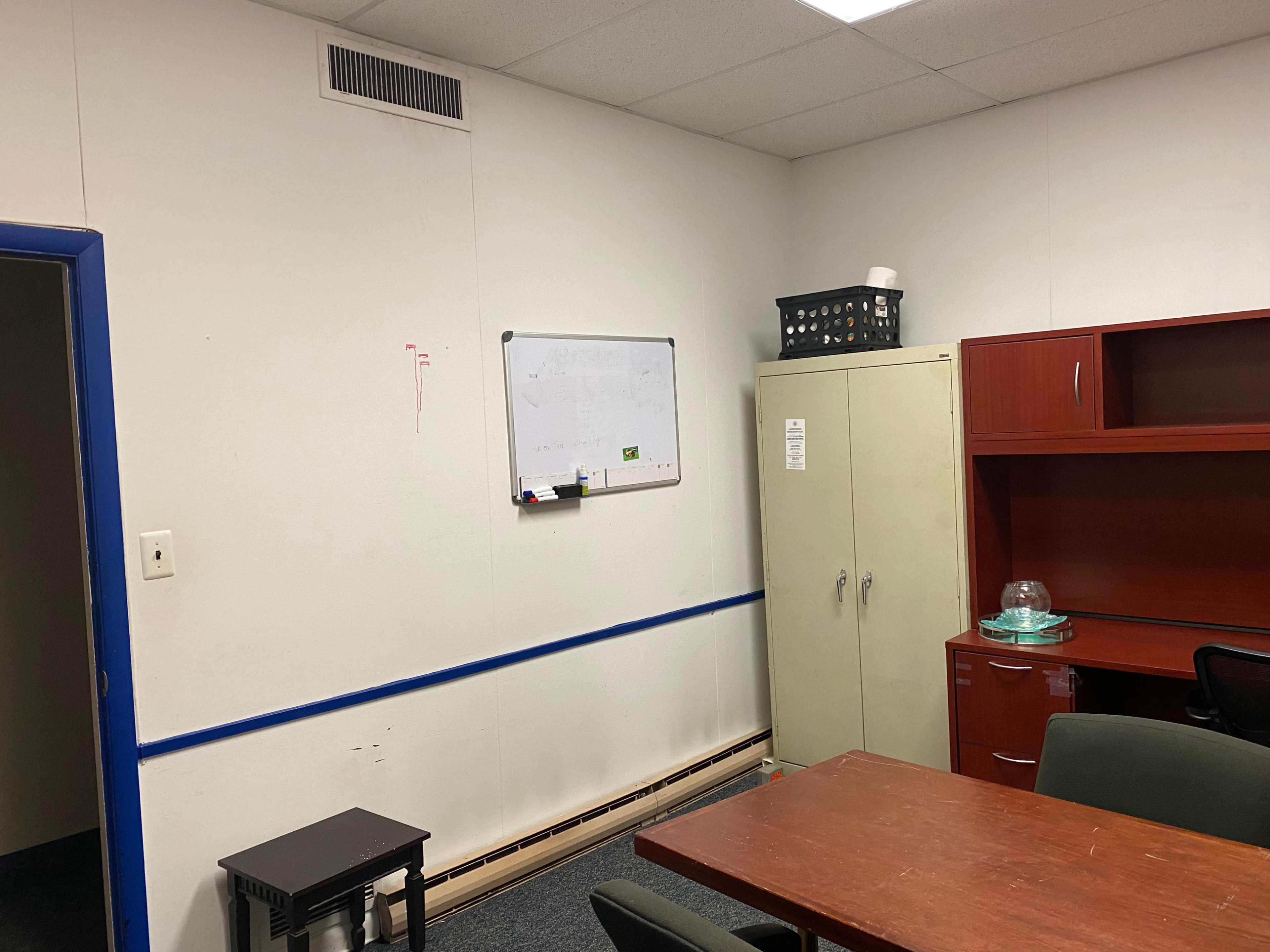 The image shows a small office space with a table, chairs, a whiteboard on the wall, a cabinet, and a shelf with a basket.