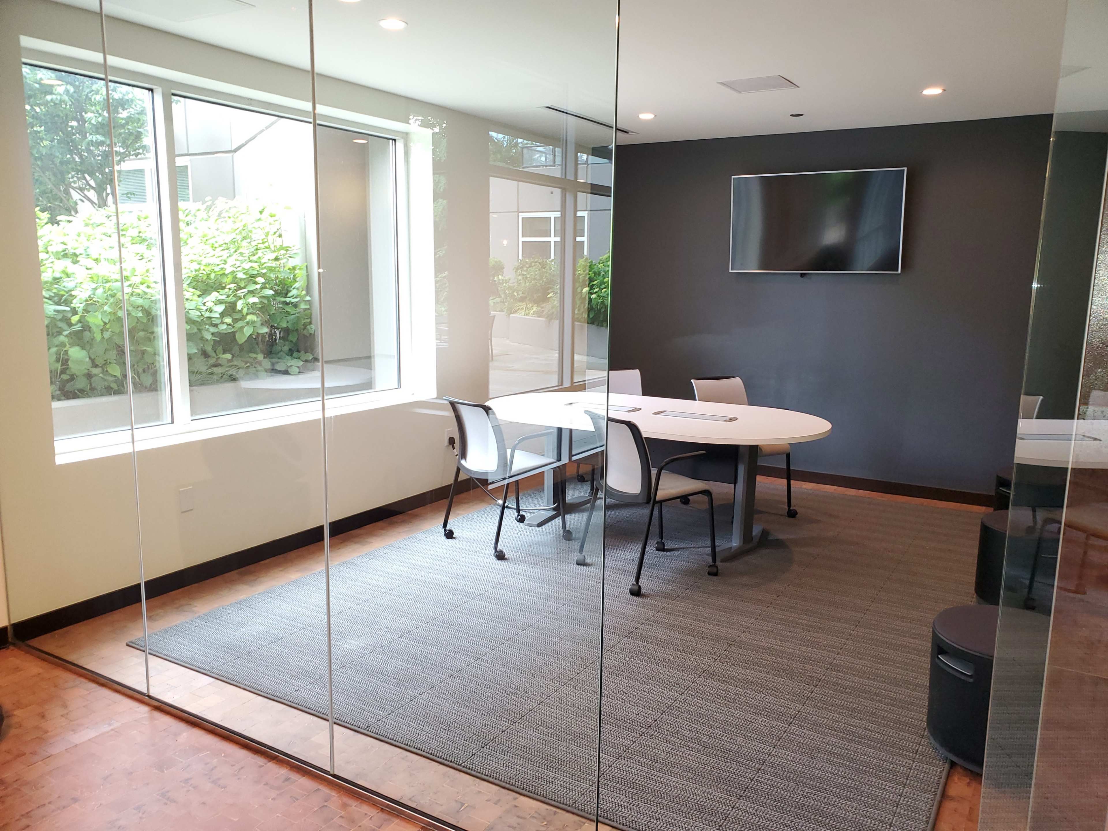 A modern conference room with a round table, four chairs, a large television on the wall, and a green view outside through a glass wall.