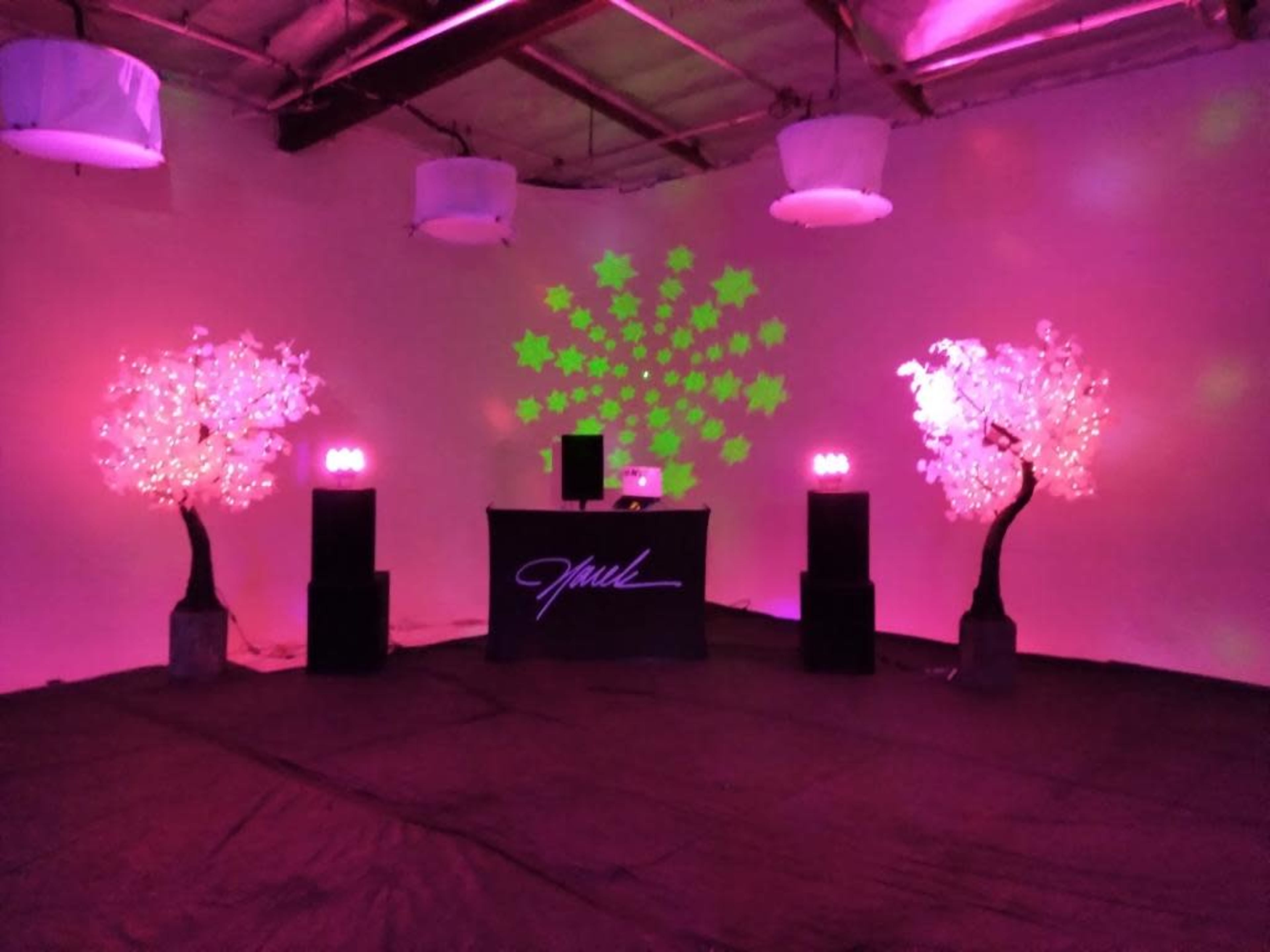 An event space featuring two illuminated trees, a DJ booth with a laptop, and speakers set against a wall displaying star-shaped light projections.