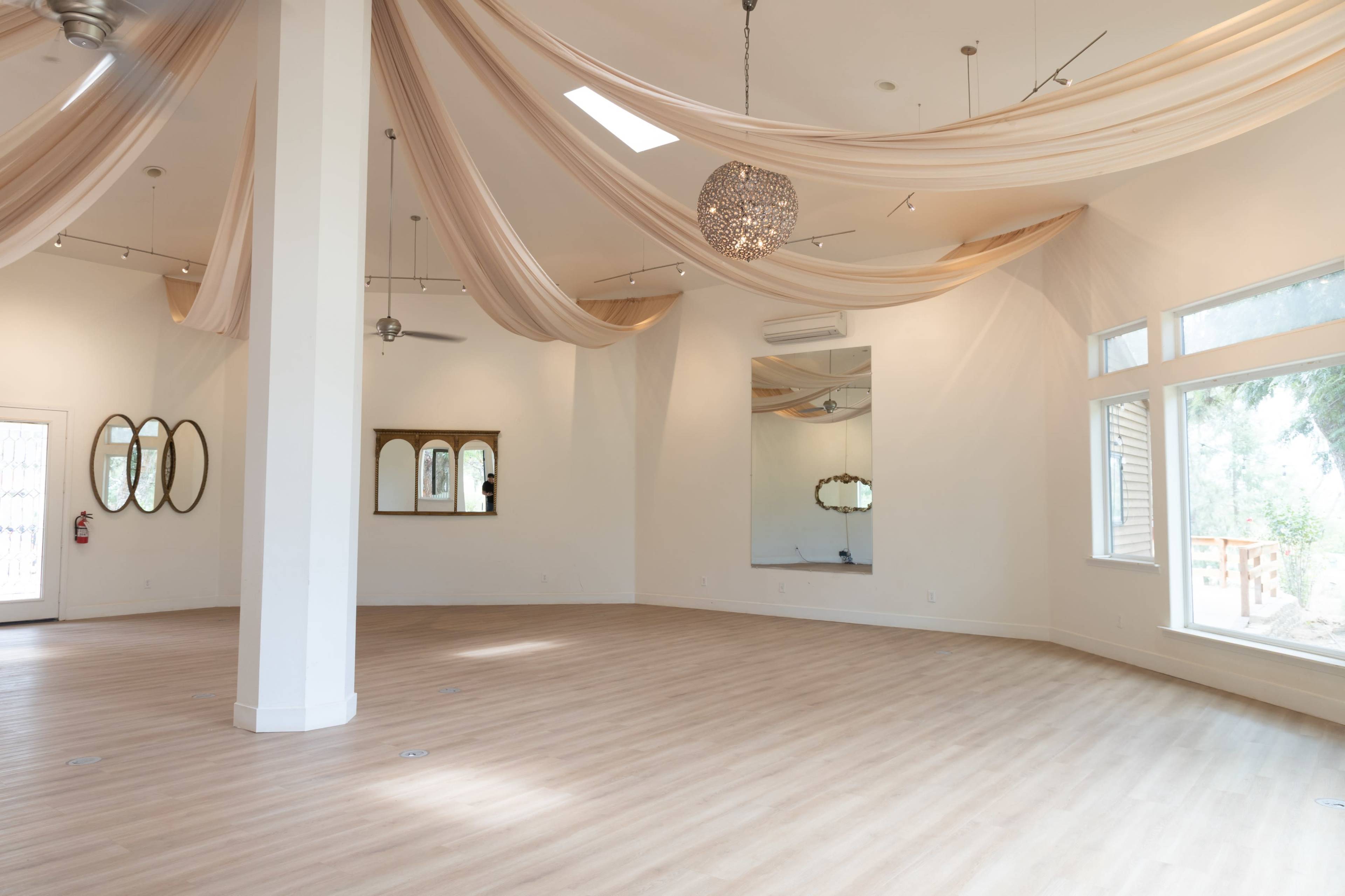The image shows a spacious, brightly lit room with light wooden flooring, adorned with draped fabric on the ceiling and a chandelier hanging down.