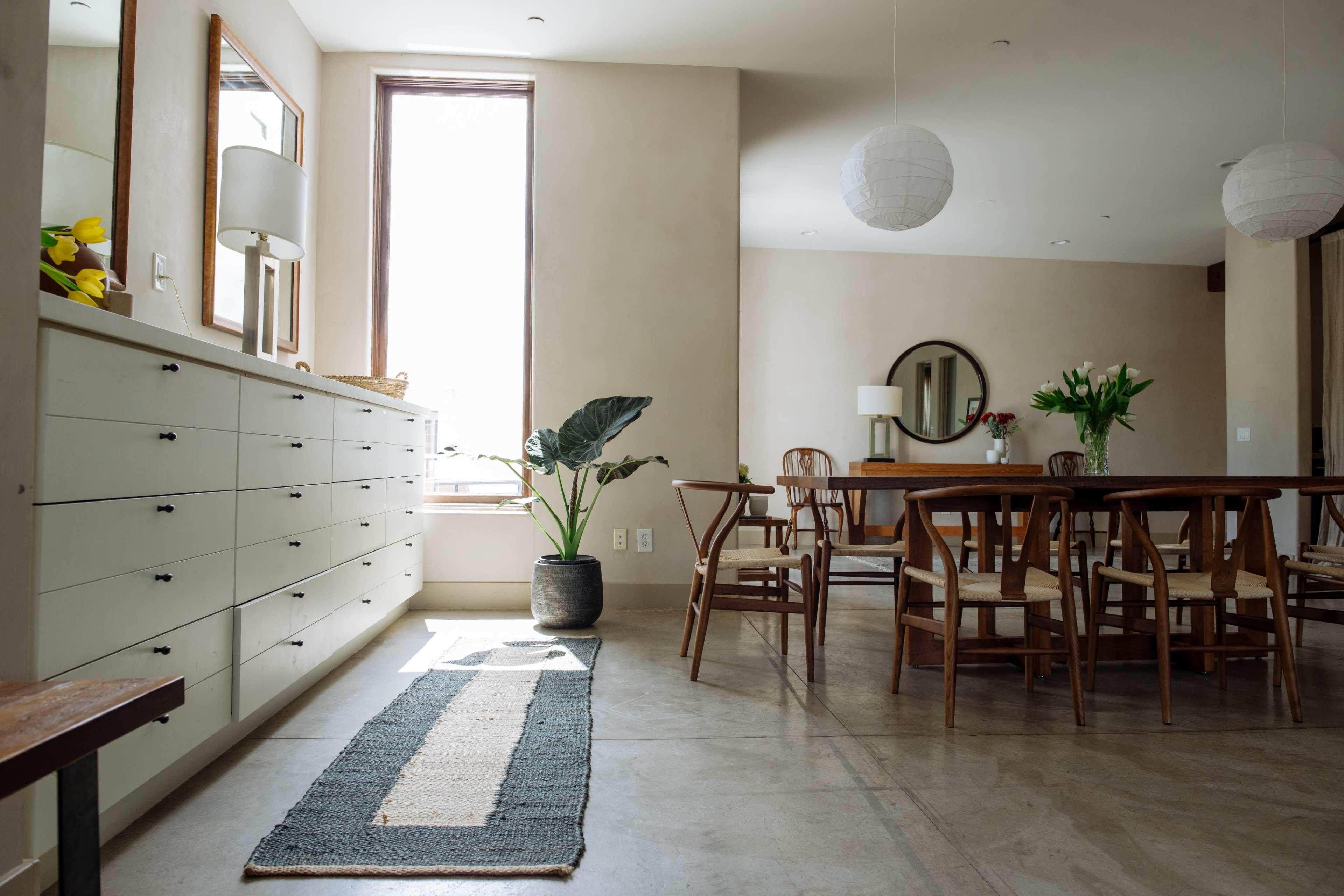 The image shows a modern interior space featuring a dining area with a wooden table, chairs, and a light-colored dresser, illuminated by natural light from a large window.