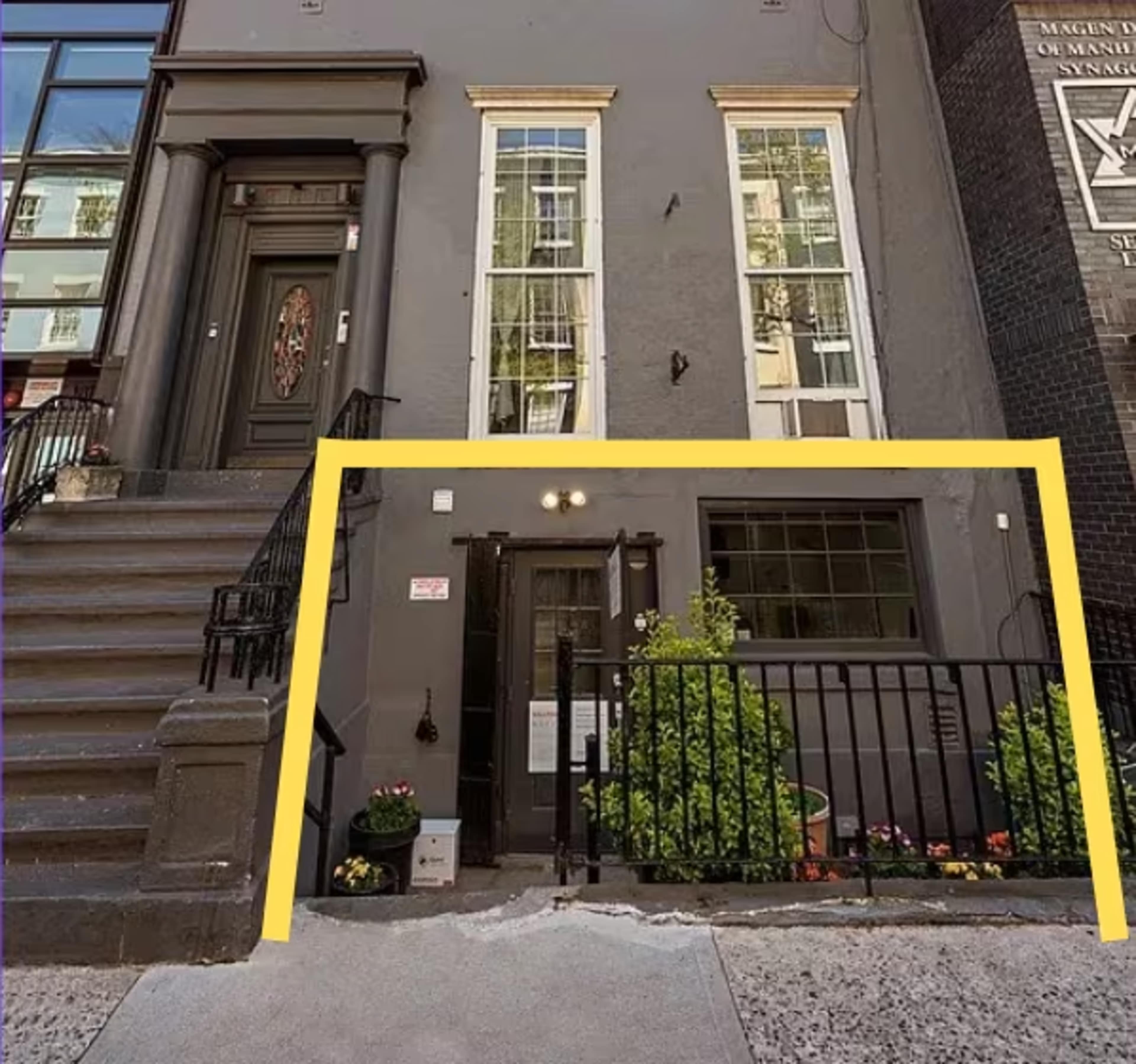 The image shows a gray building with two front entrances, one featuring a decorative door and the other a smaller entrance with a railing and planters.