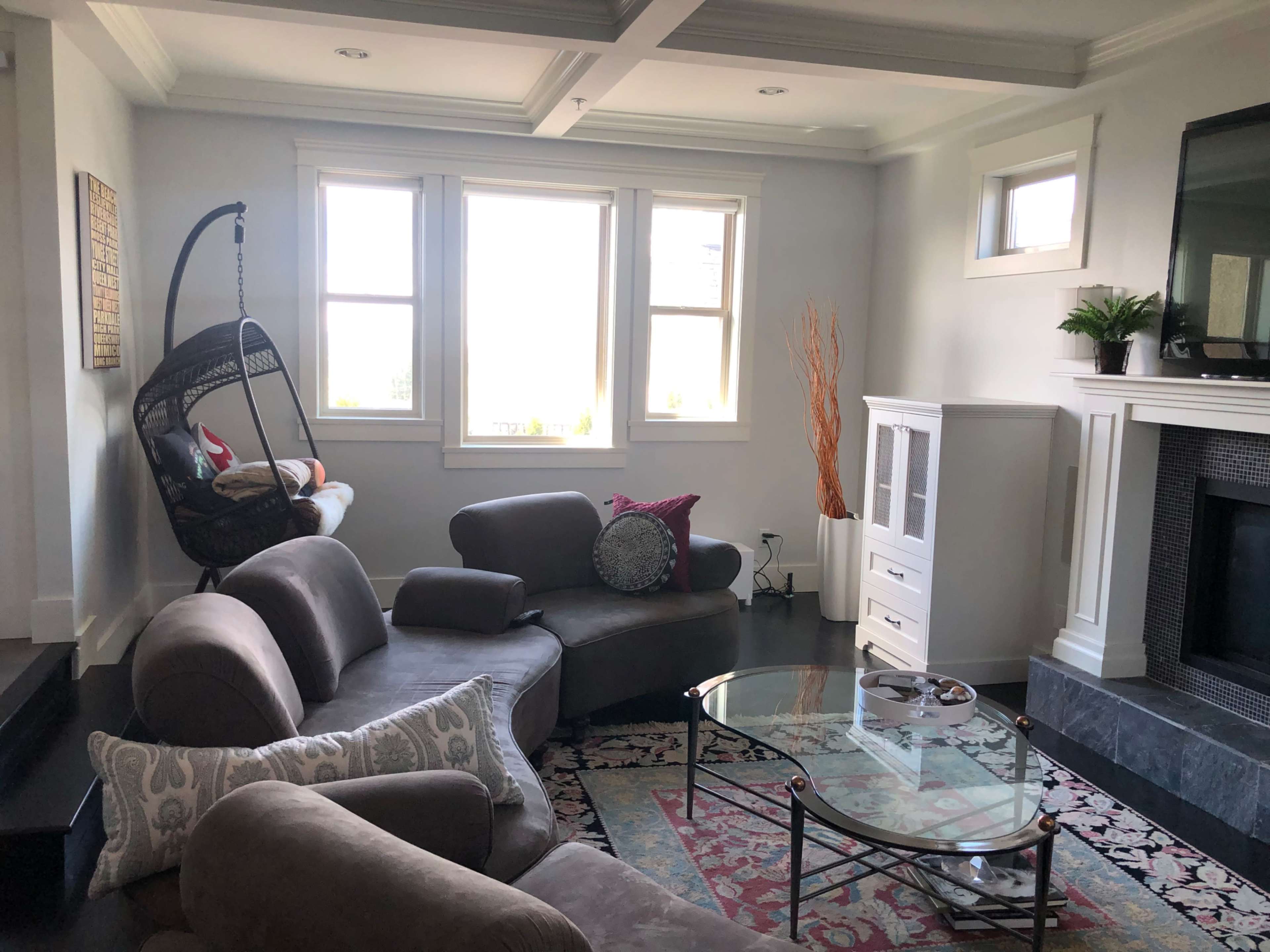 The living room features a curved gray sectional sofa, a glass coffee table, and large windows that allow natural light to enter the space.
