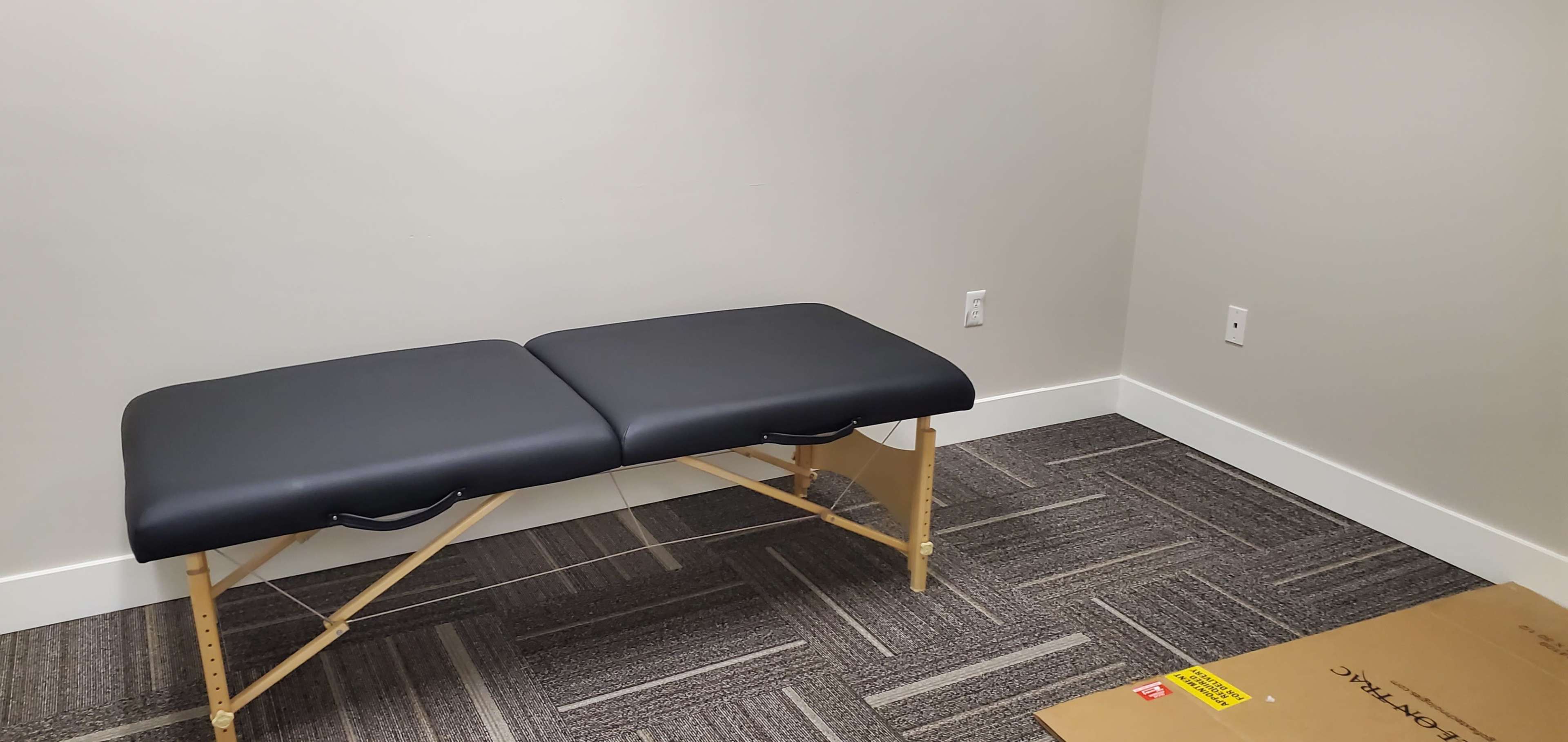 A black massage table is positioned against a plain gray wall in a sparsely furnished room with carpeted flooring.