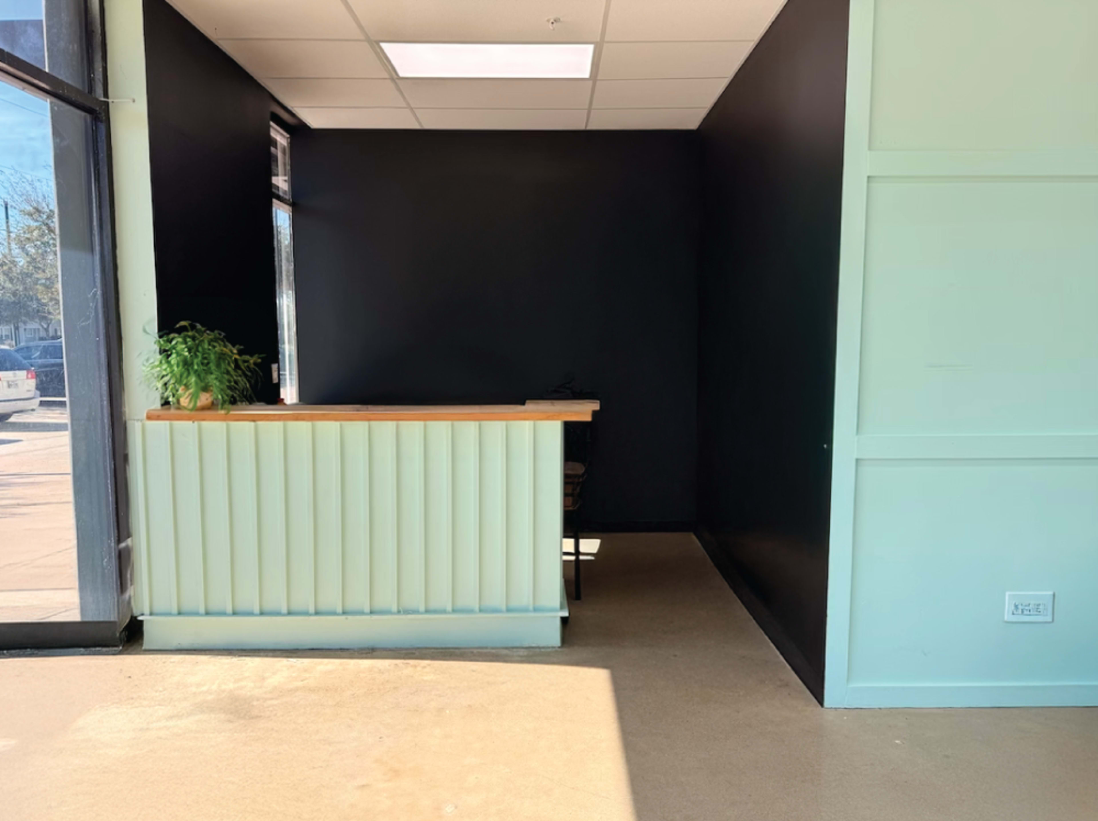 The image shows an empty reception area with a light green counter and a dark wall, illuminated by natural light from a large window.