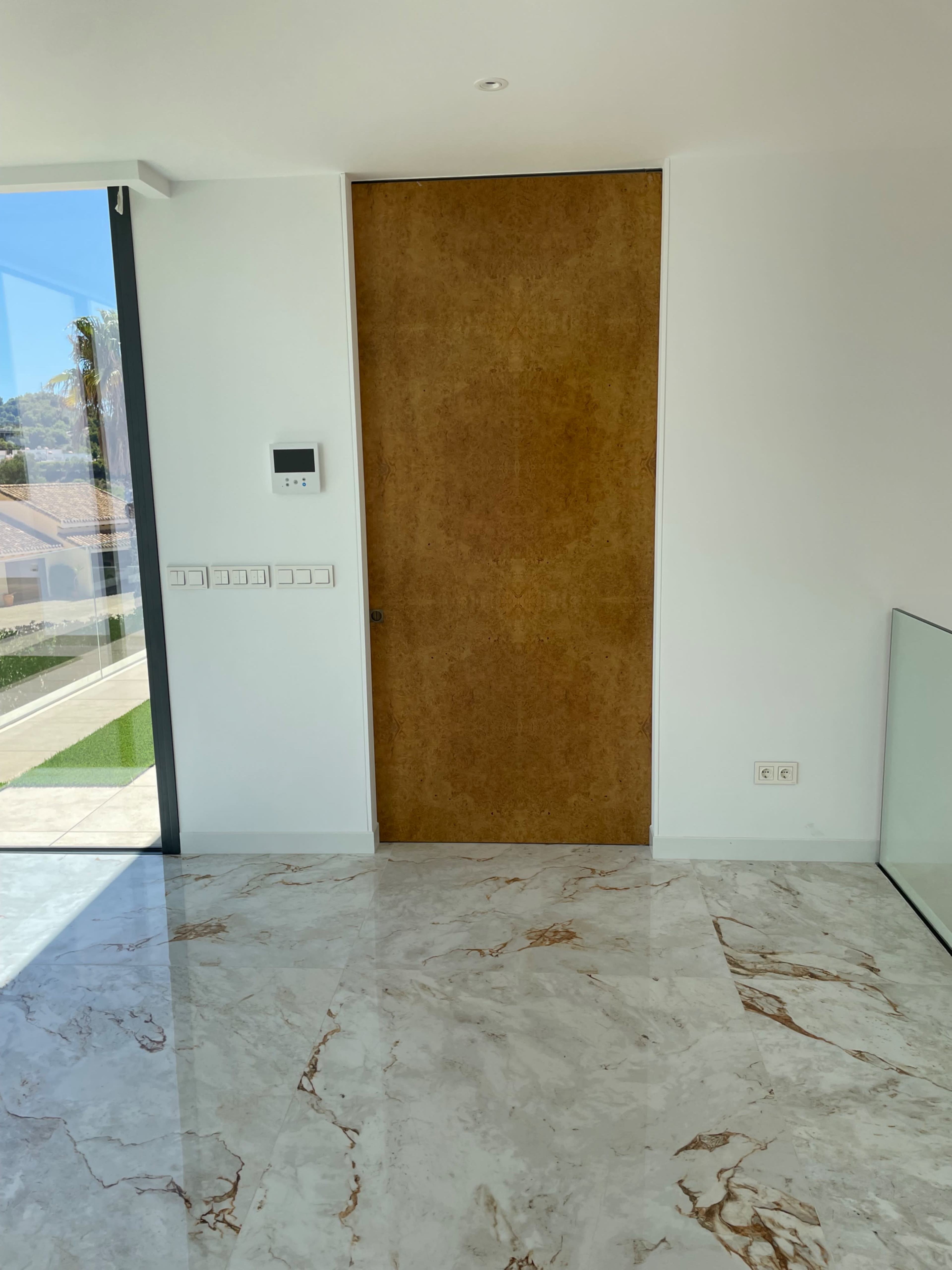 A wooden door is situated in a minimalist room with marble flooring and large windows letting in natural light.