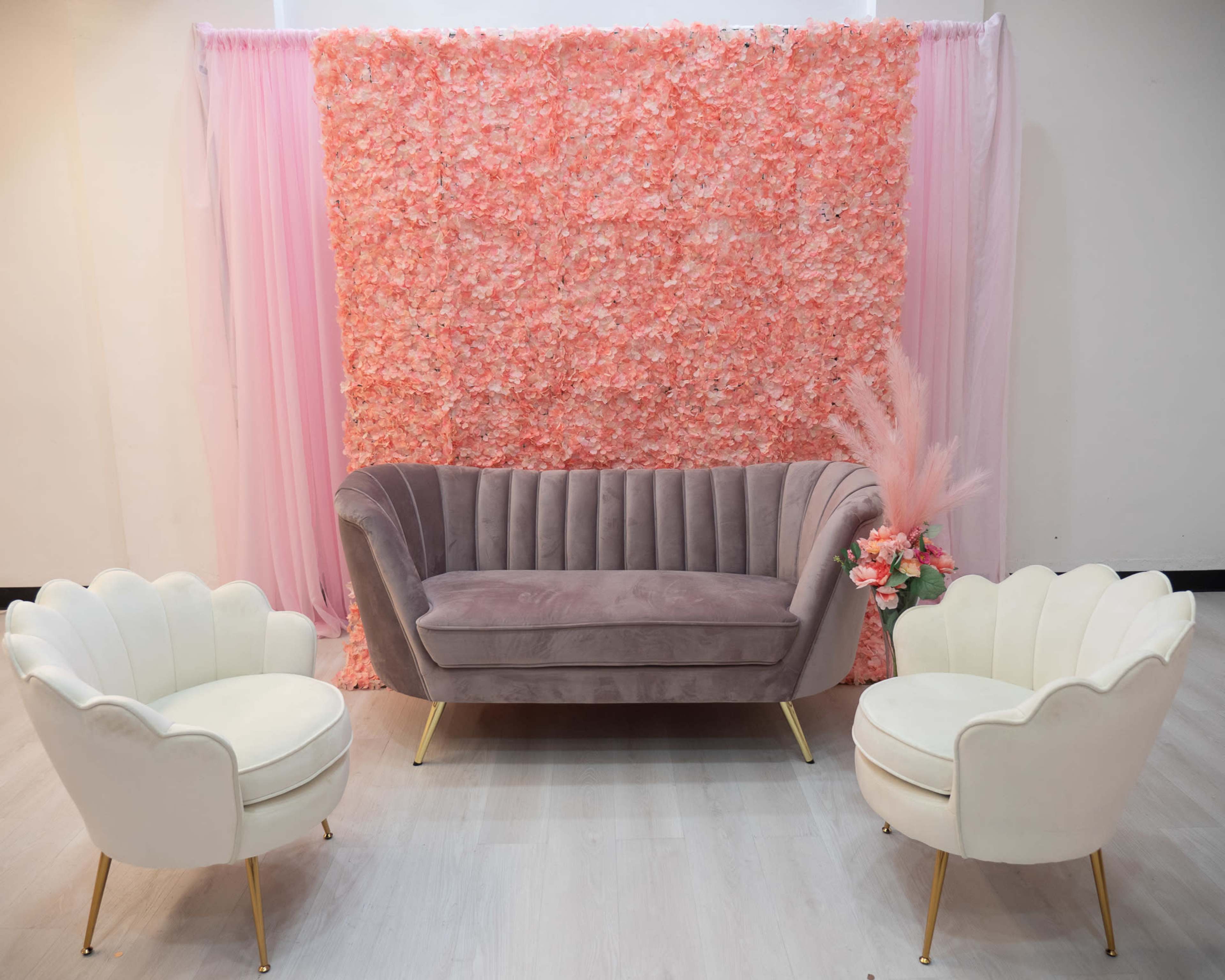 A modern seating arrangement featuring a grey velvet sofa and two white armchairs, set against a floral pink backdrop.