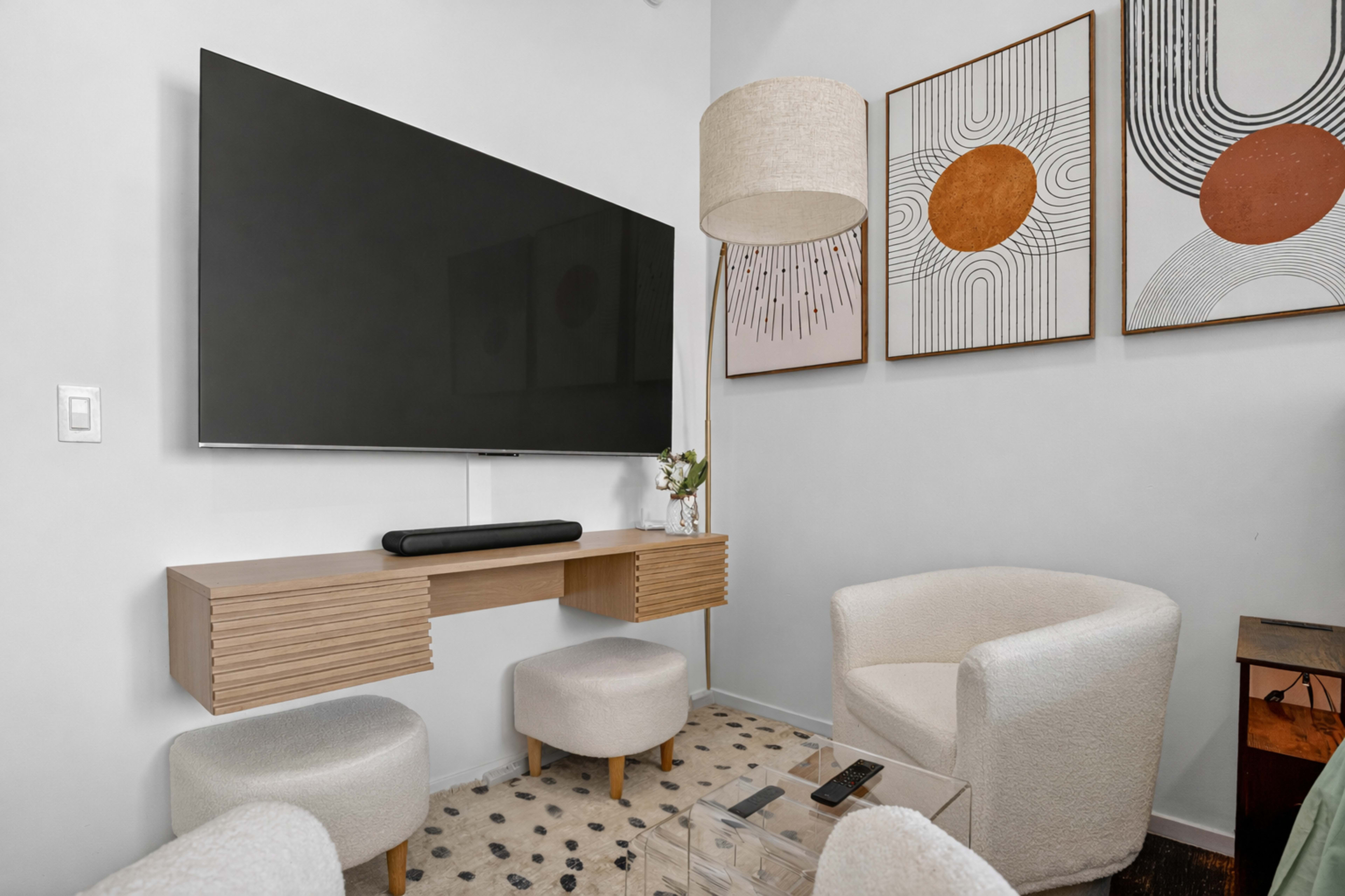 The image shows a modern sitting area featuring a large flat-screen TV mounted on the wall above a wooden console, accompanied by two circular stools and a white armchair, with abstract art on the walls.