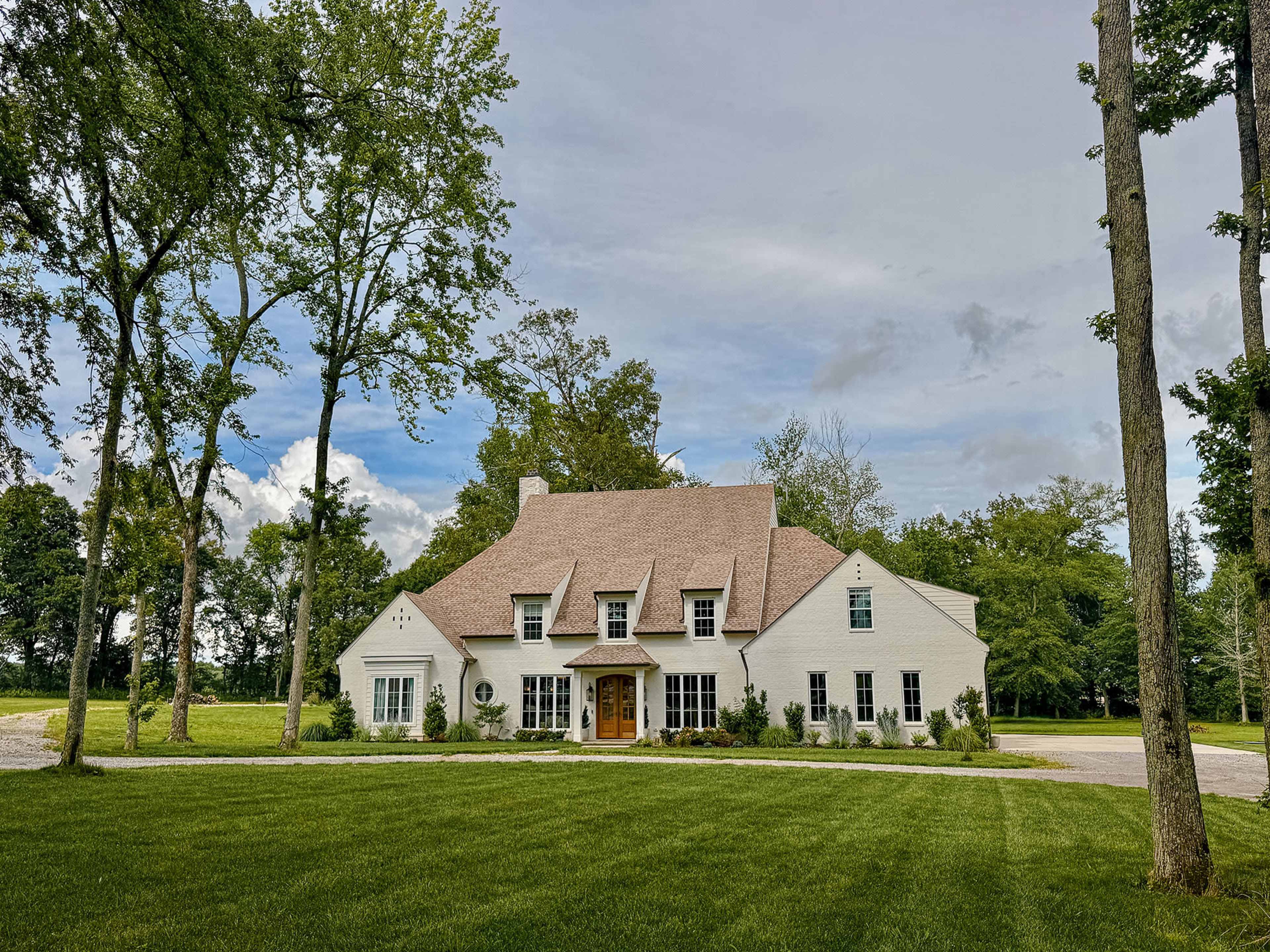 A large white house with a steeply pitched roof is surrounded by trees and a green lawn.
