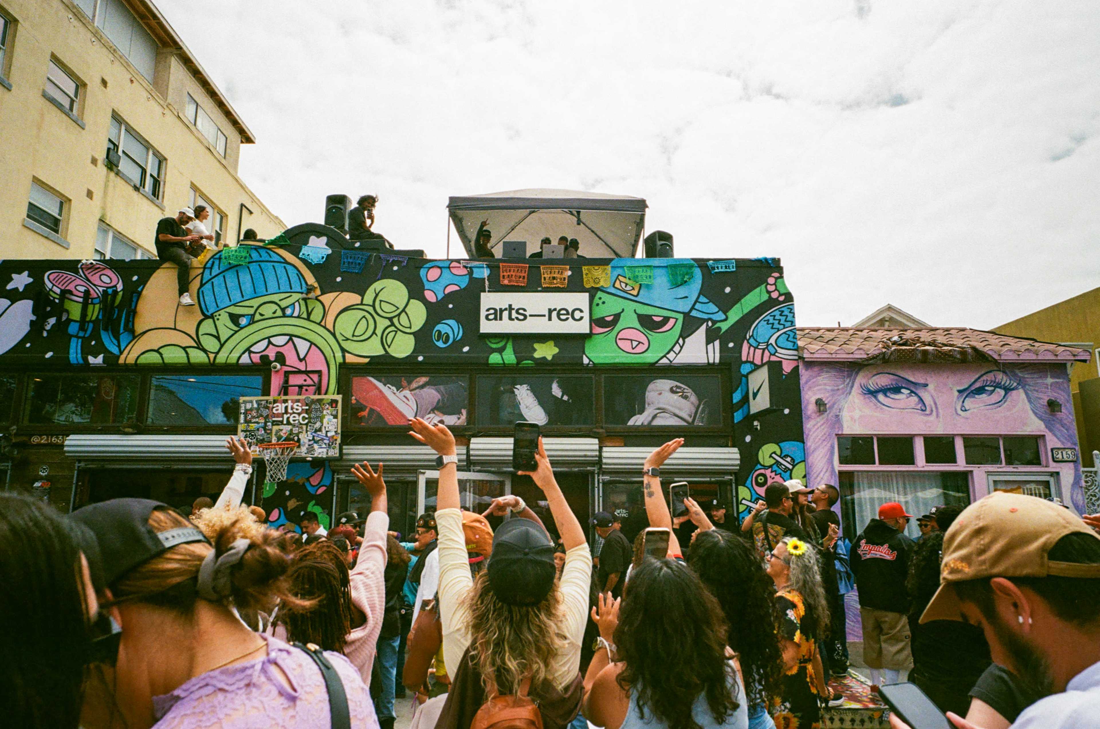 A crowd gathers in front of a colorful building adorned with graffiti, while performers are on a stage above, with people raising their hands in excitement.